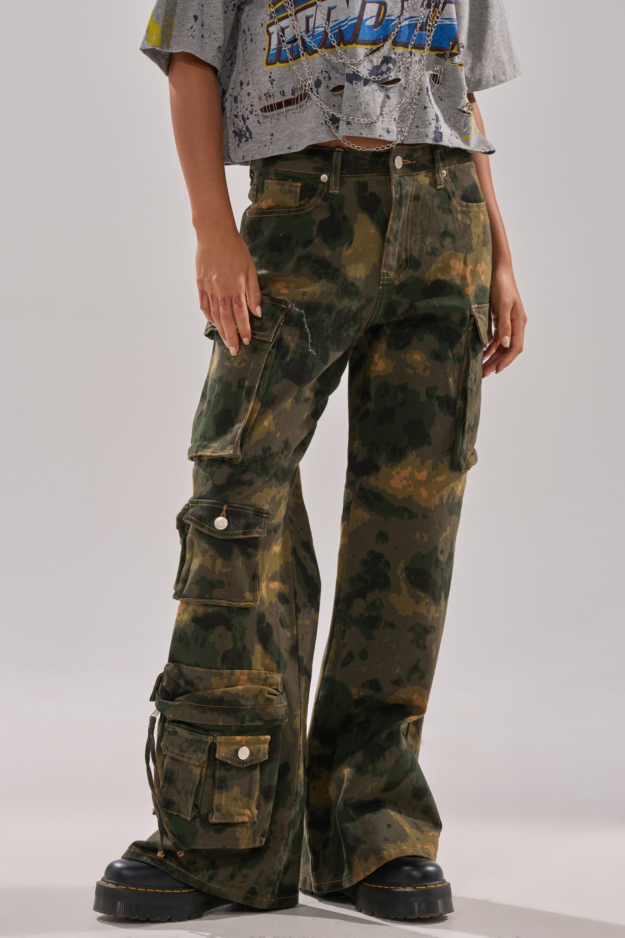 A person wearing the WASHED UP CARGO CAMO PANT with utility pockets, paired with a distressed cropped graphic tee and black platform shoes. 0