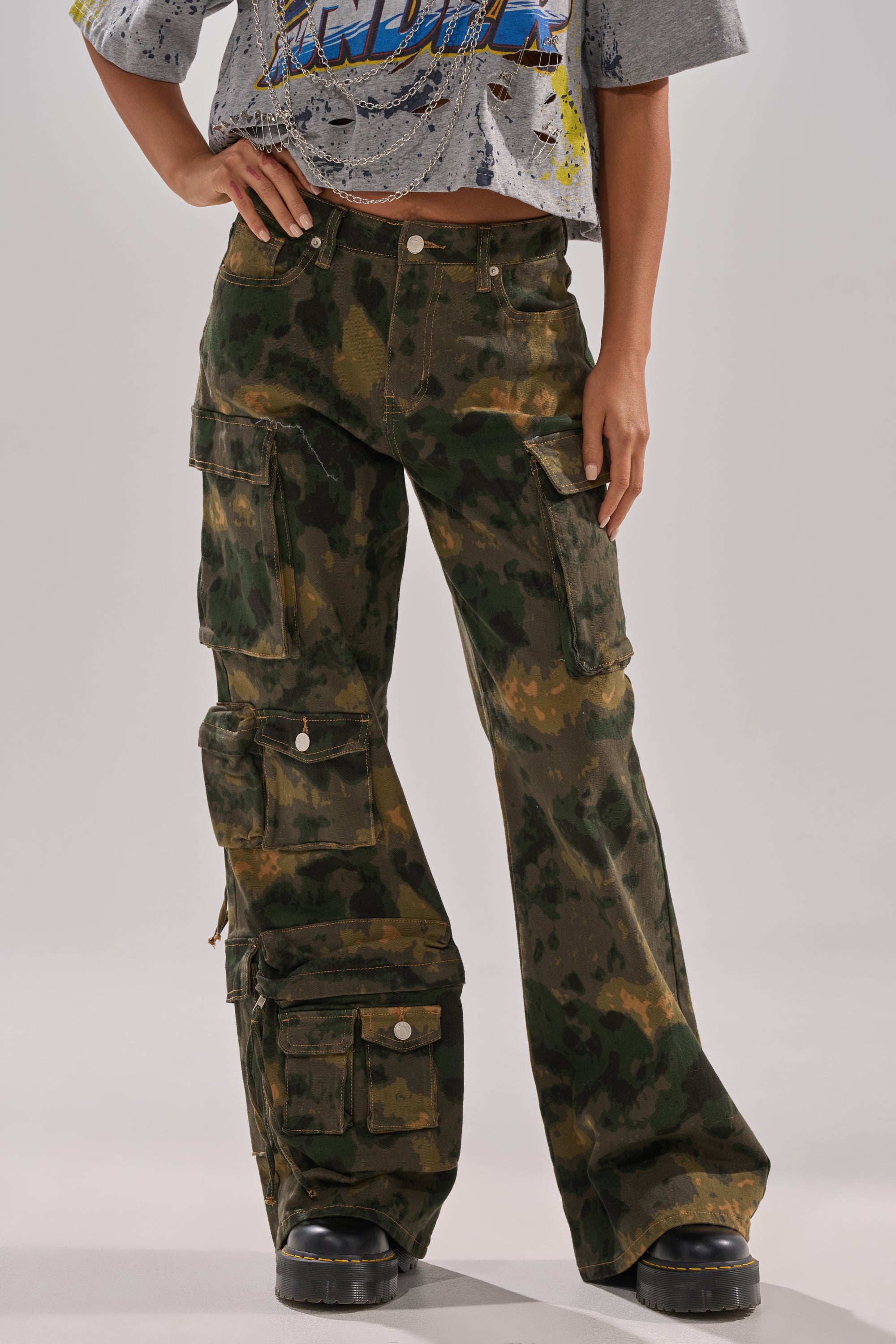 A person wears the WASHED UP CARGO CAMO PANT with utility pockets and black chunky shoes, styled with a distressed crop t-shirt. The image is cropped at the shoulders. 1