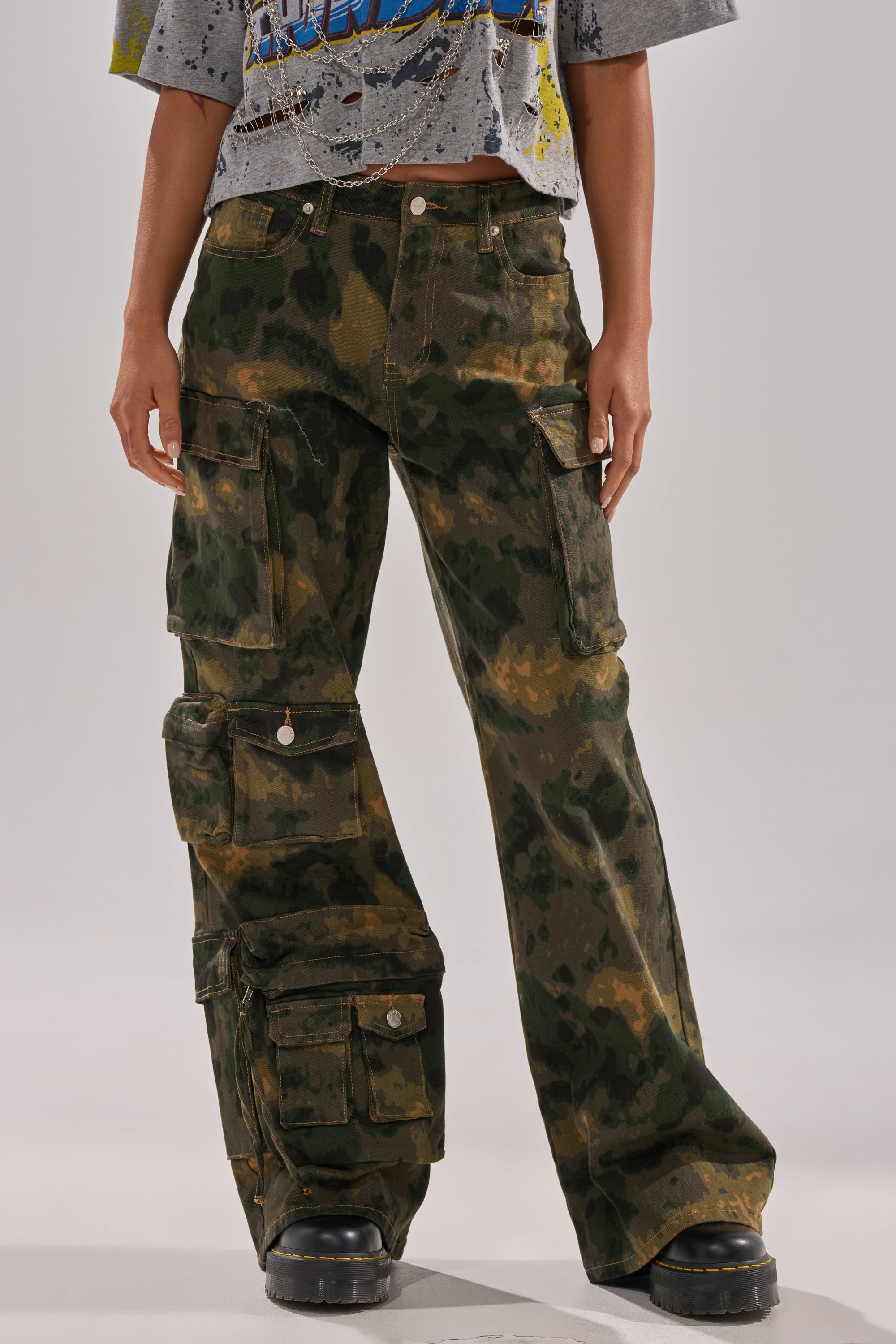 Person wears WASHED UP CARGO CAMO PANT—loose-fitting, green with utility pockets—paired with chunky black shoes and a cut-off graphic distressed tee. The face is not visible. 2