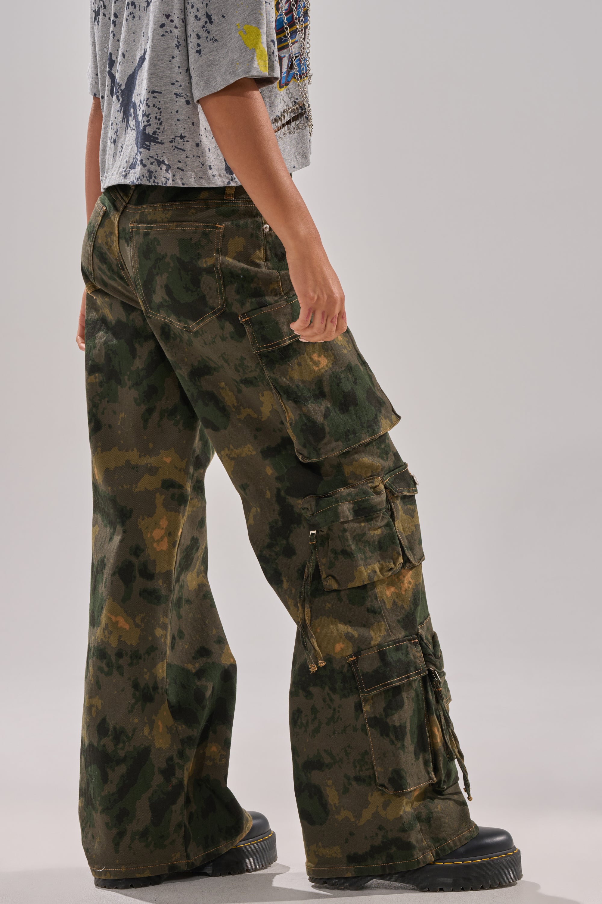 A person stands sideways wearing the WASHED UP CARGO CAMO PANT with utility pockets, black platform shoes, and a gray short-sleeved printed shirt; their upper body is partially visible. 3
