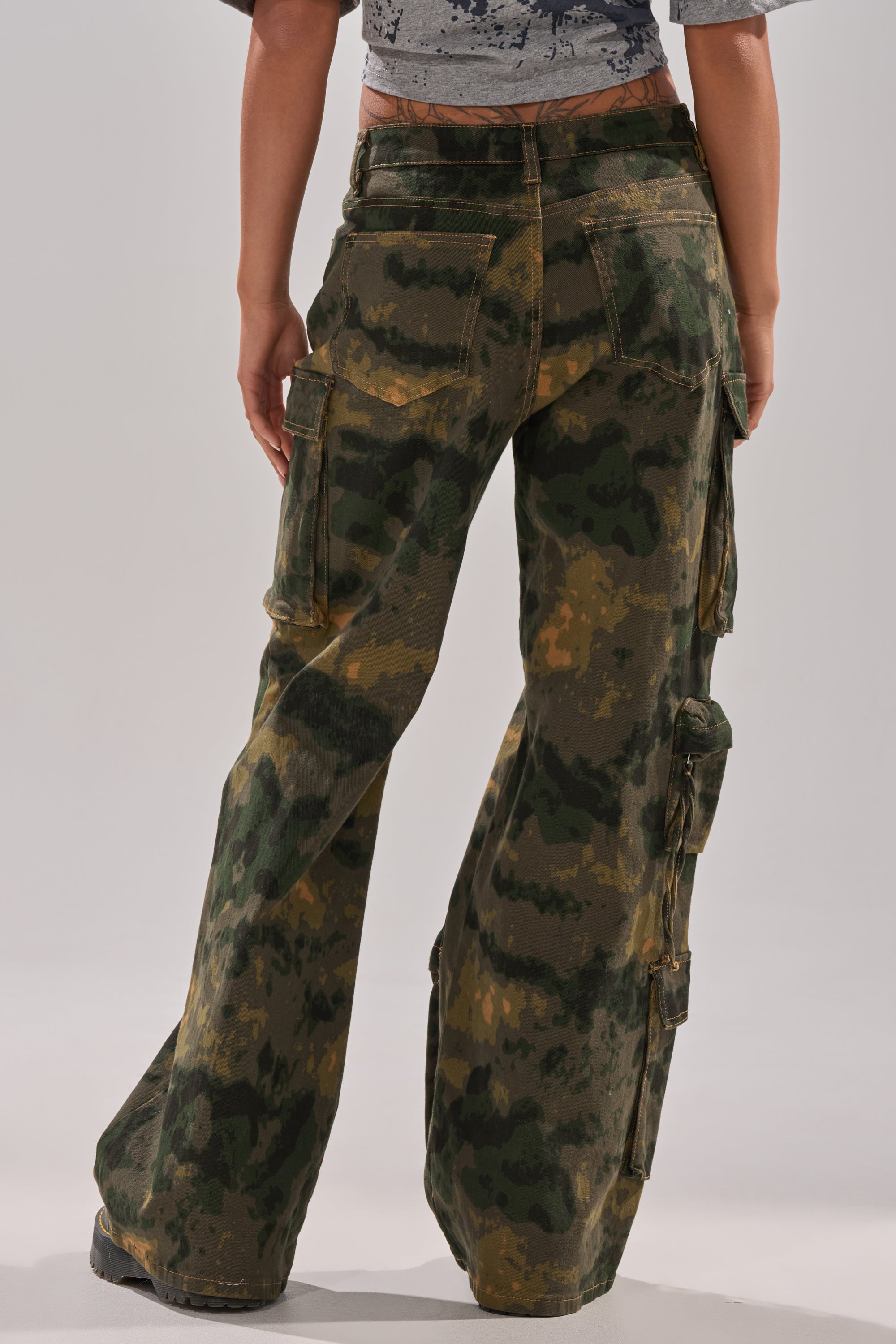 A person faces away wearing the WASHED UP CARGO CAMO PANT—loose-fitting, high-waisted cargo pants with utility pockets, featuring a green, brown, and black camo pattern and a wide-leg silhouette. The upper body is partially visible. 4