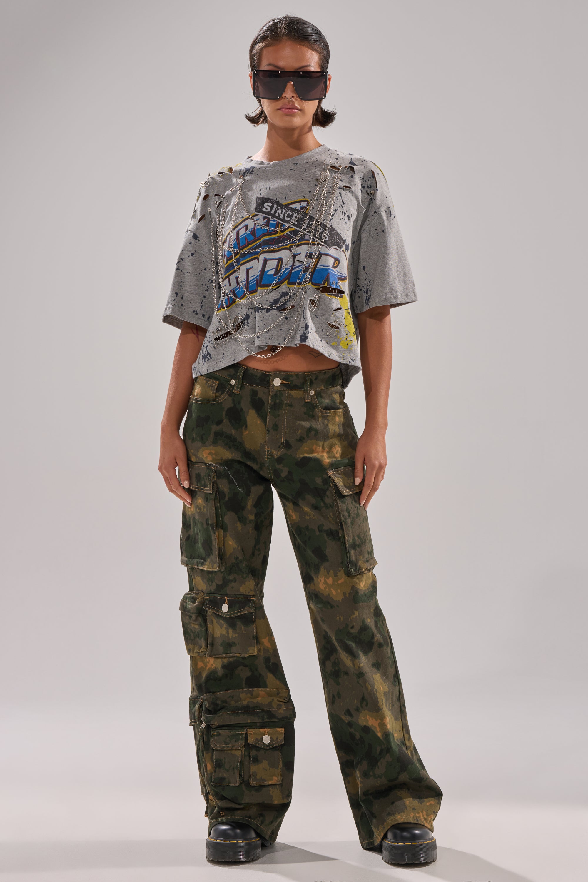 A person stands confidently with hands in pockets, wearing large sunglasses, a distressed gray graphic tee, and the WASHED UP CARGO CAMO PANT—loose-fitting with a wide-leg silhouette—against a plain light background. 5