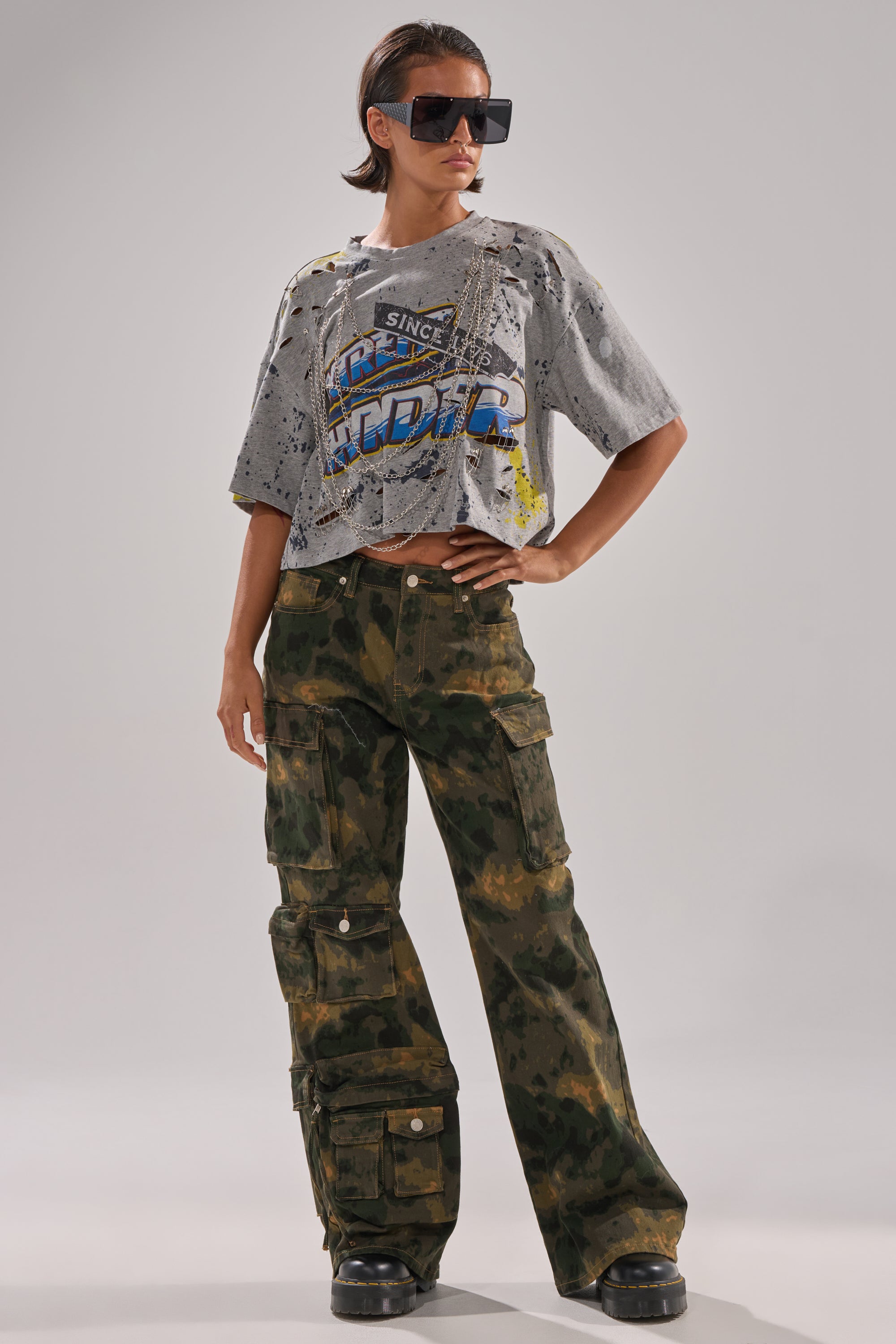 A person poses confidently against a plain background wearing oversized sunglasses, a distressed gray graphic crop top, and the WASHED UP CARGO CAMO PANT featuring utility compartments and a wide-leg silhouette. 6