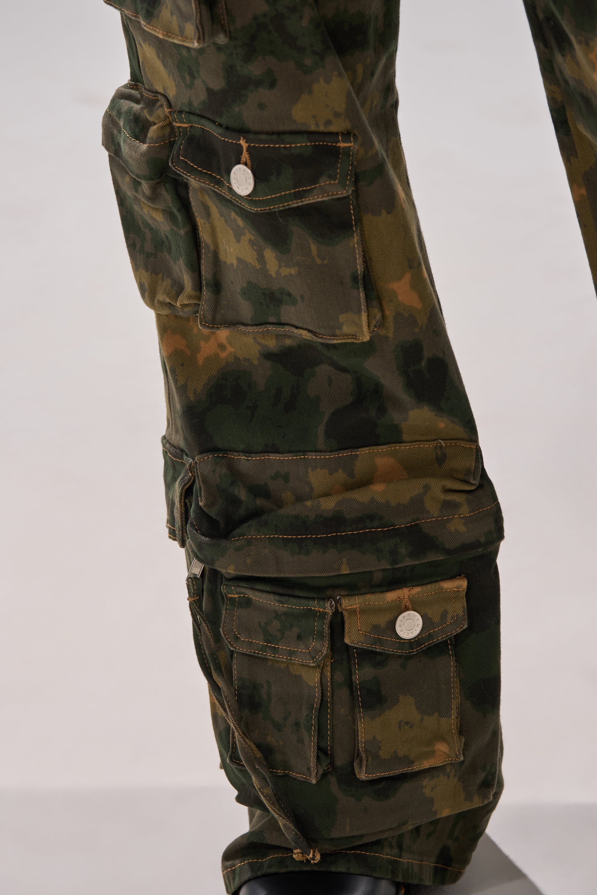 Close-up of the WASHED UP CARGO CAMO PANT in a wide-leg fit, showing multiple utility cargo pockets with metal button closures in green, brown, and black camo patterns. Lower leg and part of a black shoe visible. 8