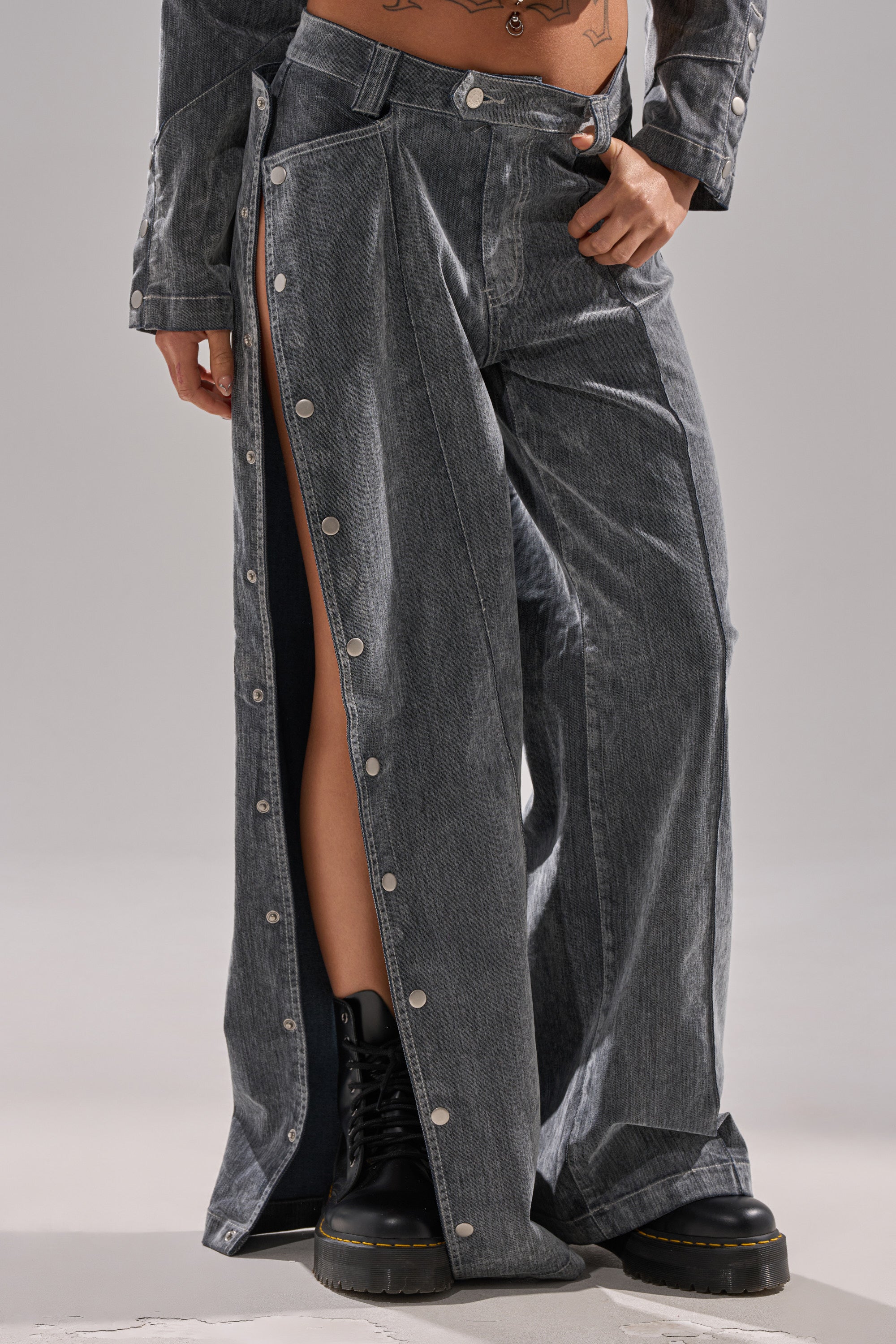 A person wears the CATCHING EYES VELVET DENIM WIDE-LEG PANT in gray with side snap buttons partly open, paired with black lace-up boots. Tattoos are visible on their torso and hand.