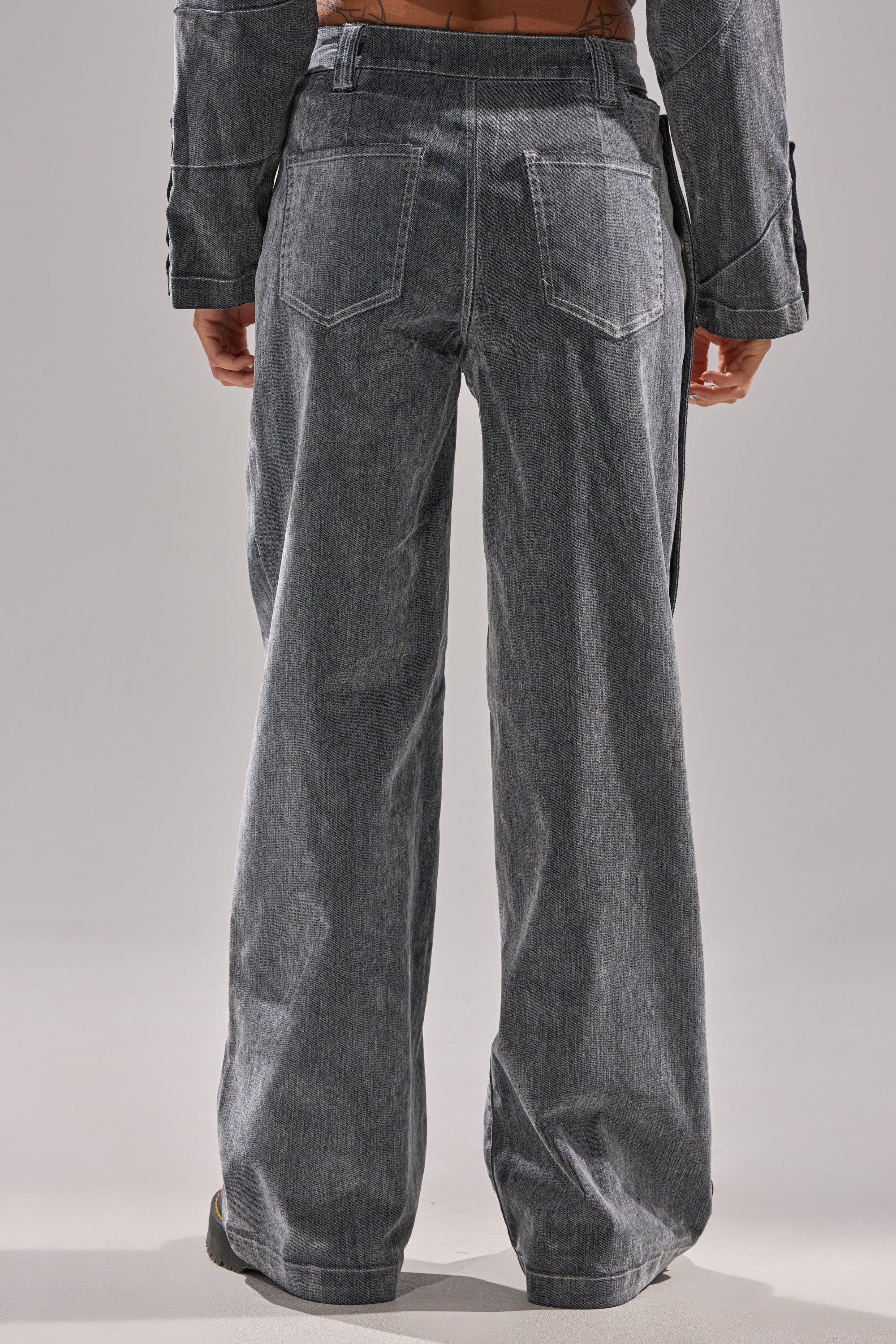 A person stands with their back to the camera, showcasing the CATCHING EYES VELVET DENIM WIDE-LEG PANT in gray, featuring visible seams, back pockets, and a loose, wide-leg fit from waist to floor.