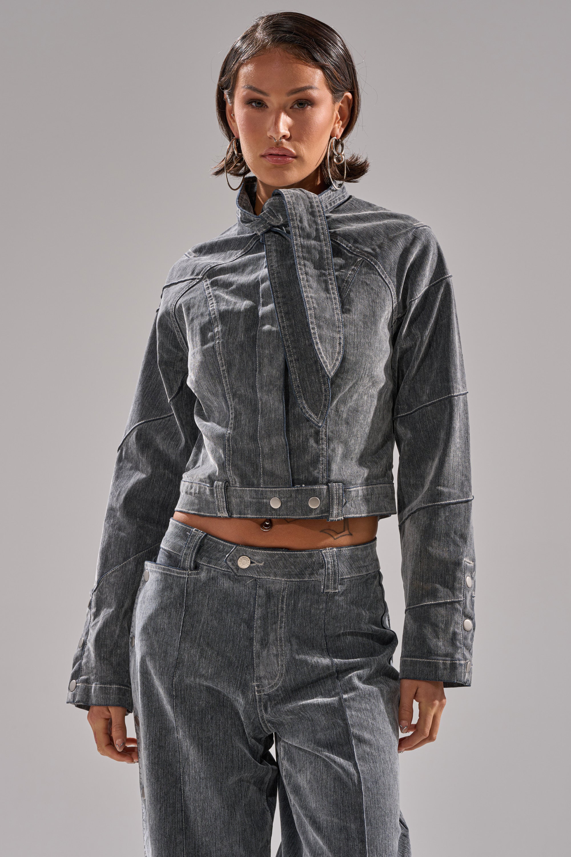 A person stands confidently in the CATCHING EYES VELVET DENIM BLOUSE— a vintage-inspired gray jacket with a high collar, metallic buttons, visible seams, and cropped fit. The plain background highlights the standout fashion piece.