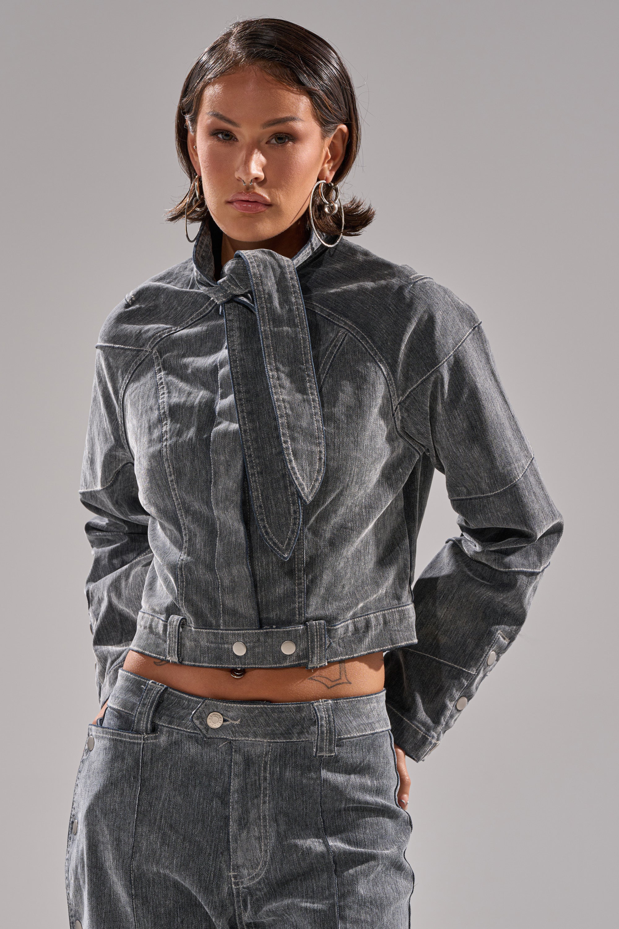 A person with short dark hair models the CATCHING EYES VELVET DENIM BLOUSE, featuring an attached tie detail, paired with matching high-waisted jeans against a plain light background.