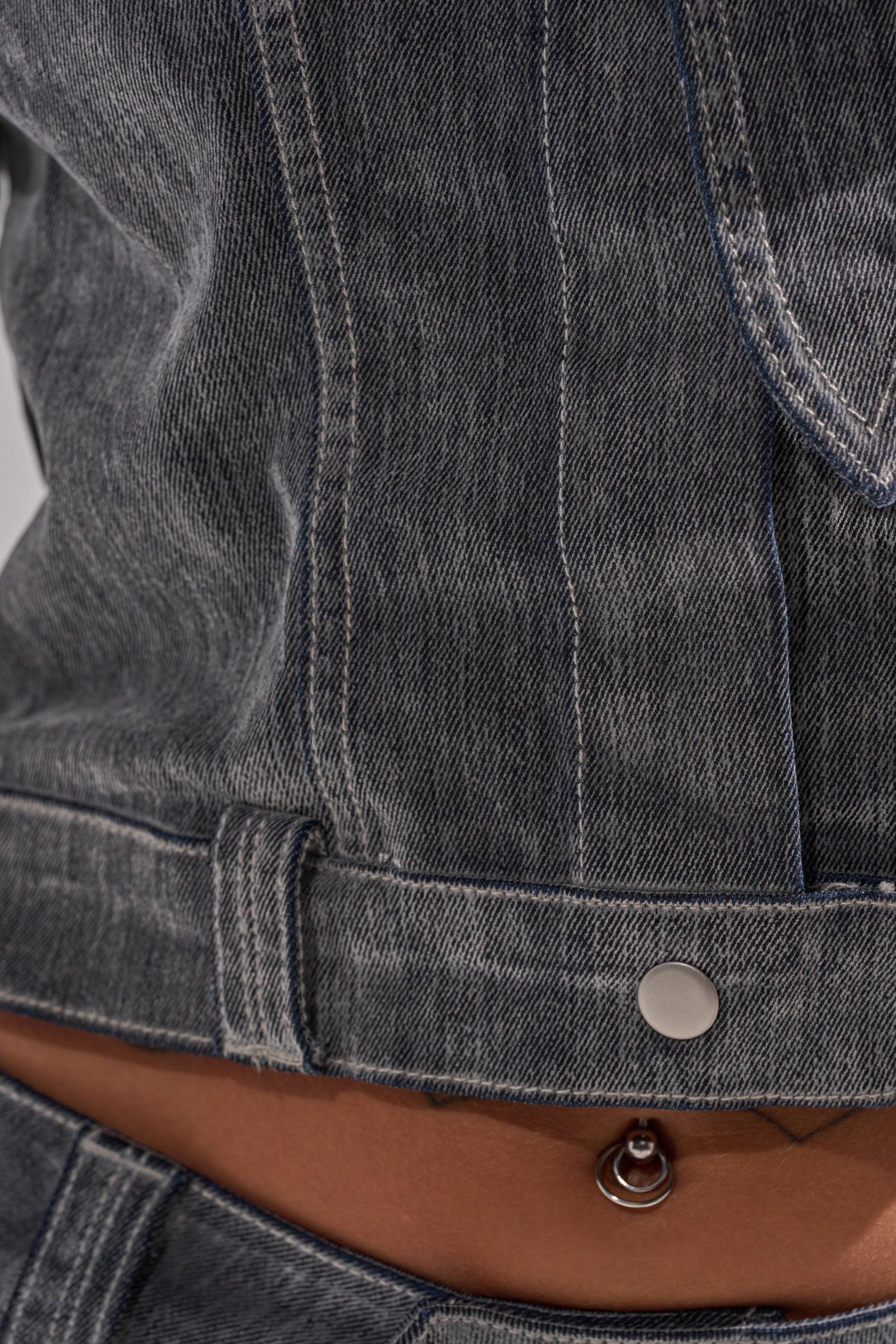 Close-up of a person wearing the CATCHING EYES VELVET DENIM BLOUSE in a faded black wash, paired with matching jeans. Their midriff shows a belly button piercing with a small charm just above the waistband.