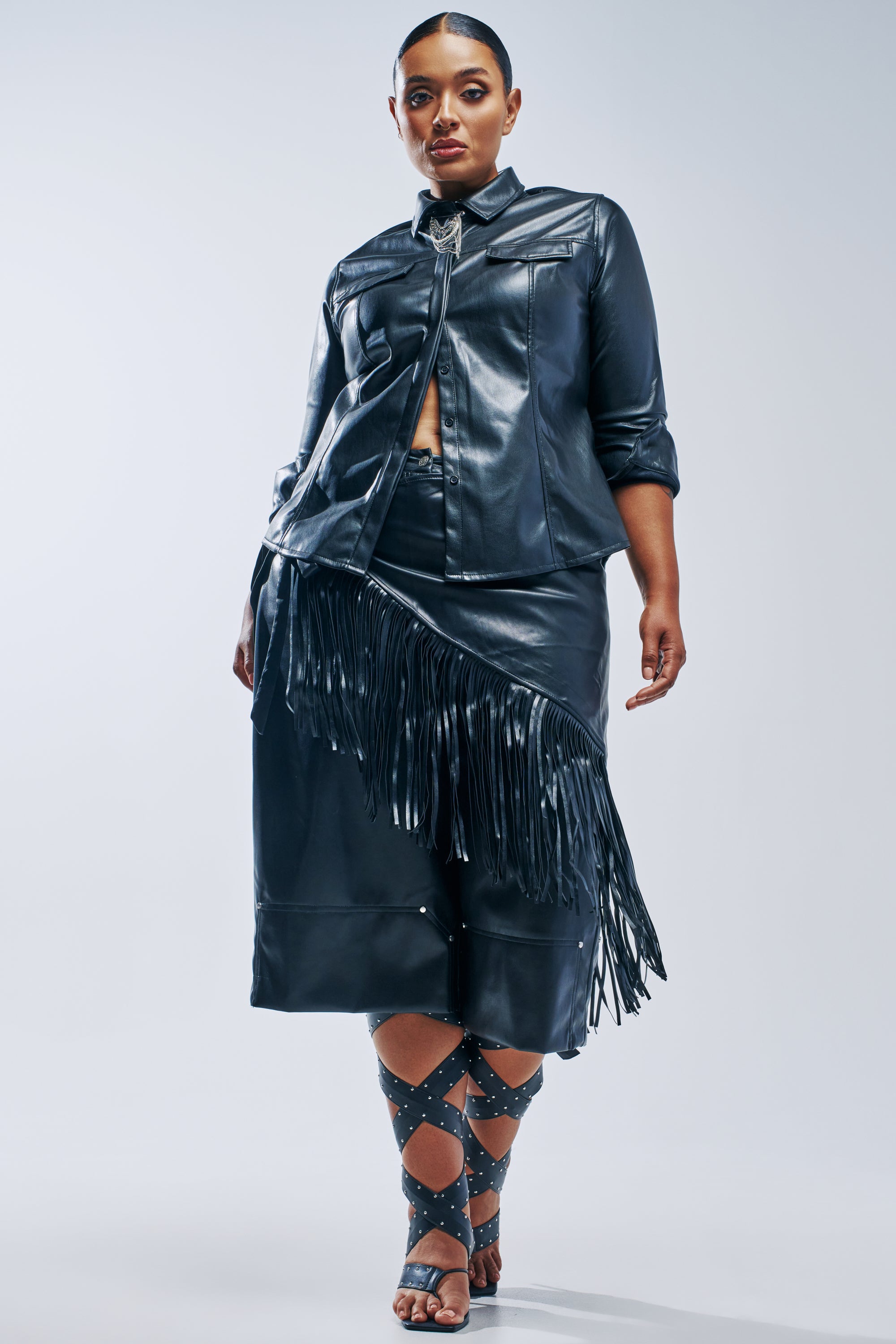 A person wearing a black faux leather button-down shirt and matching COME BACK TO ME FAUX LEATHER GAUCHO pants with fringe detailing, paired with black strappy studded sandals.