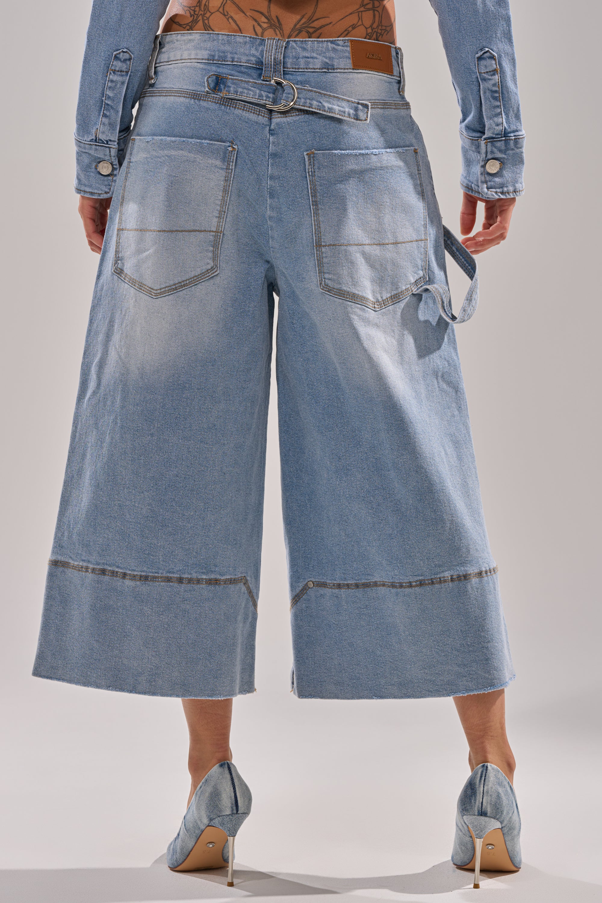 A person wears ROCK MY WORLD WIDE-LEG DENIM BERMUDA SHORTS with visible pockets and seam details, paired with matching denim heels. The cropped top of a denim jacket and a tattooed lower back are partly visible.
