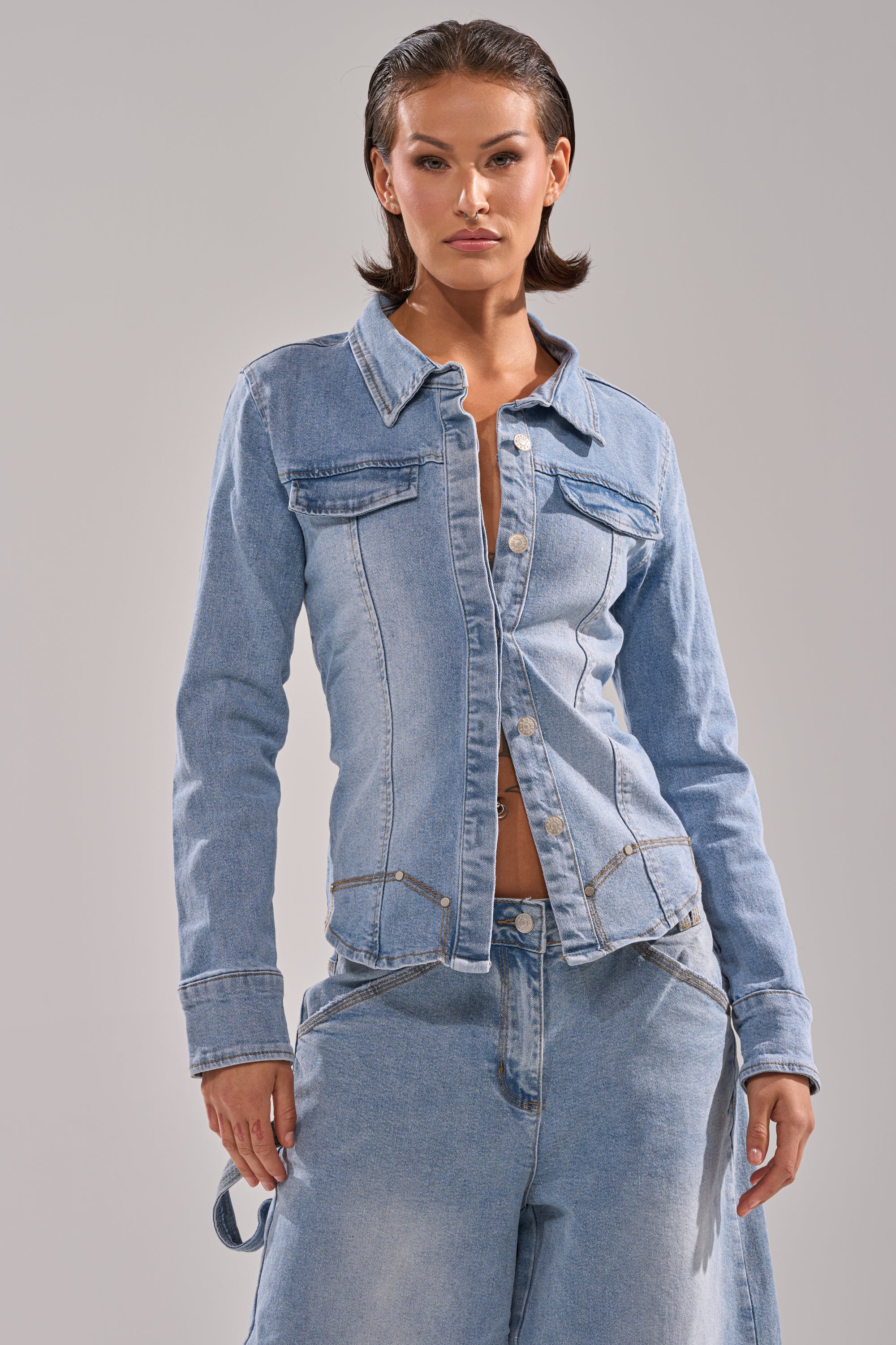 Wearing the ROCK MY WORLD DENIM BUTTON DOWN over a bare midriff with matching baggy denim jeans, a person with short dark hair stands against a plain light background.