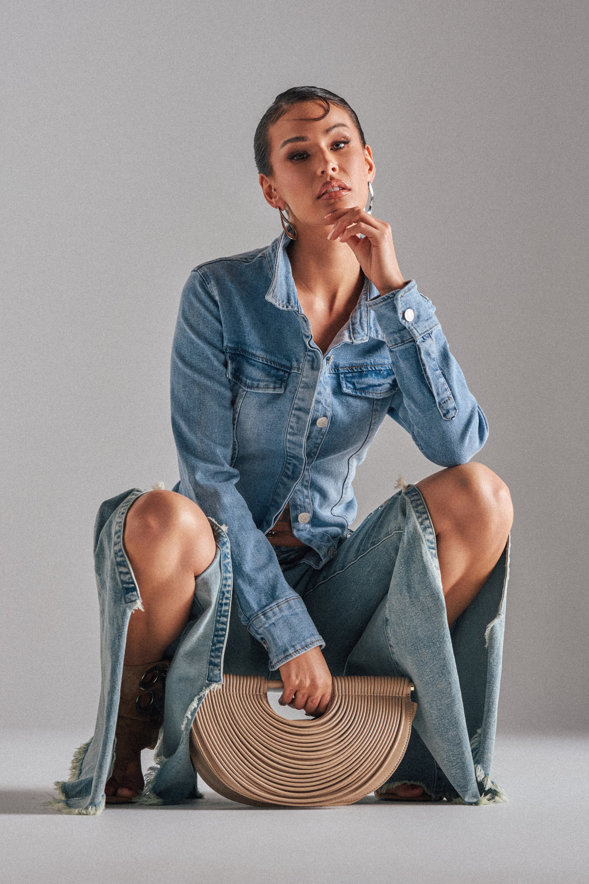 Wearing the ROCK MY WORLD DENIM BUTTON DOWN and ripped wide-leg jeans, a woman poses confidently with a beige round handbag, one hand on her chin and the other holding the bag, against a plain gray background. 1