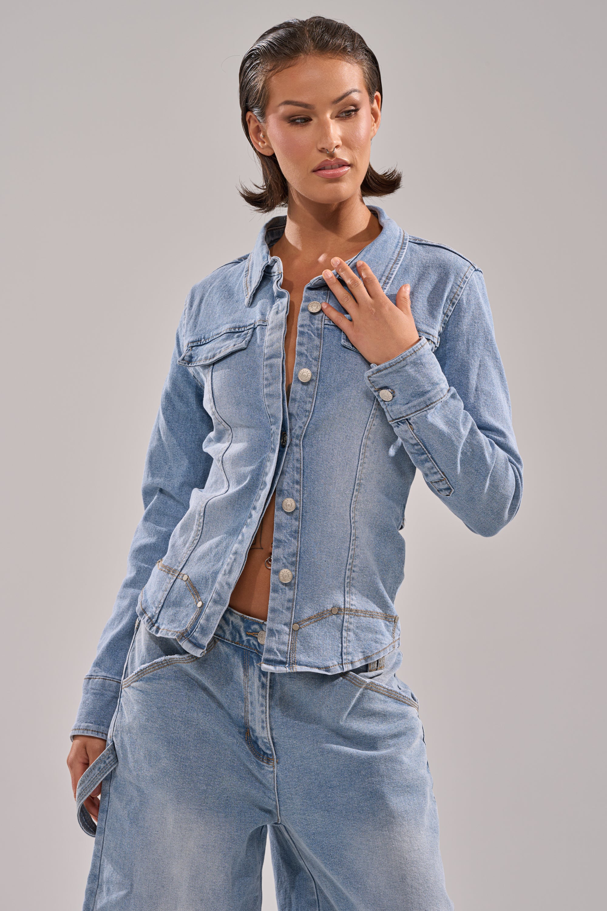 A woman wears the ROCK MY WORLD DENIM BUTTON DOWN in light blue with matching wide-leg jeans, posing with one hand on her chest and the other in her pocket against a plain light grey background.