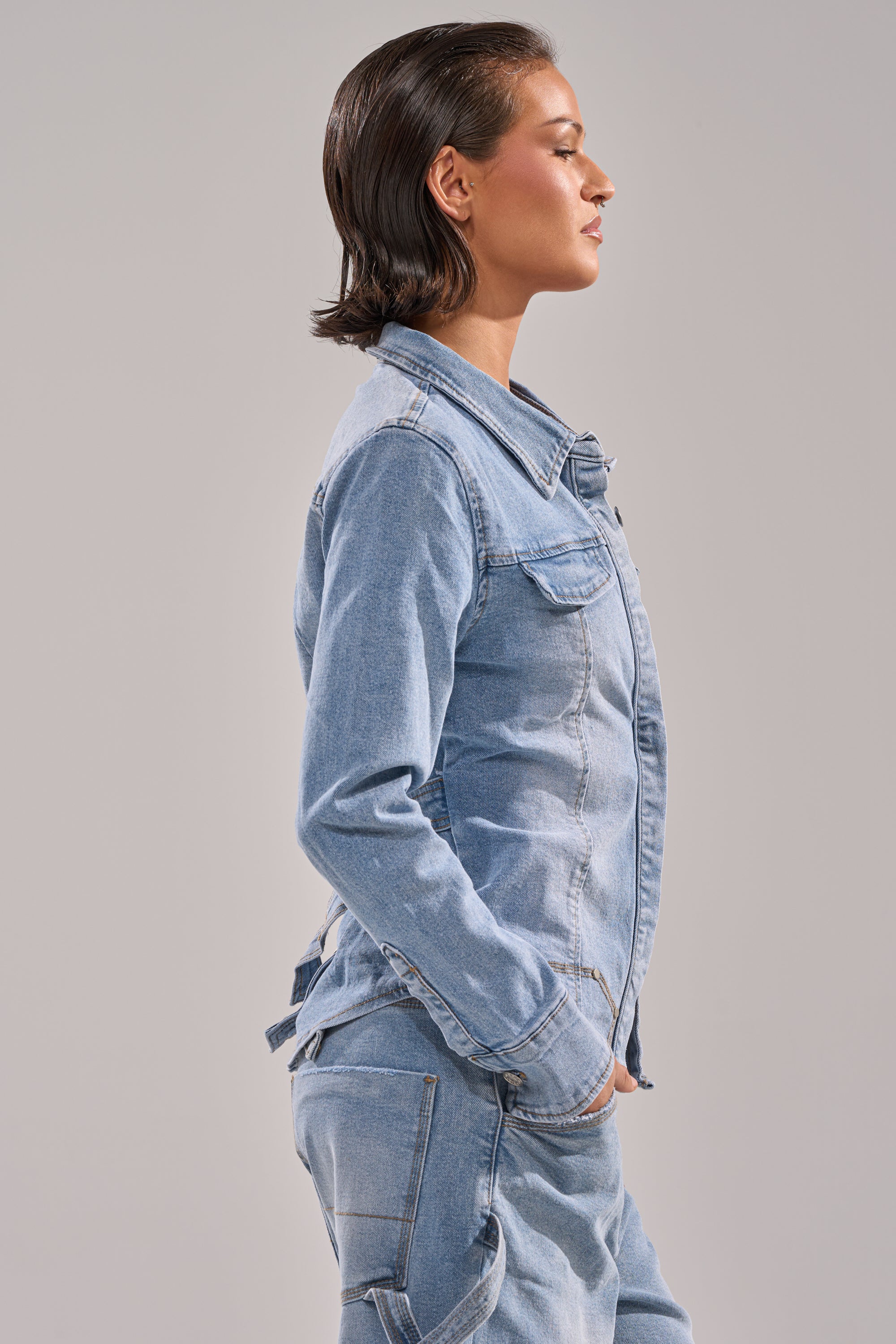 A person with slicked-back, short brown hair stands in profile with hands in pockets, wearing the ROCK MY WORLD DENIM BUTTON DOWN and jeans, against a plain light gray background.