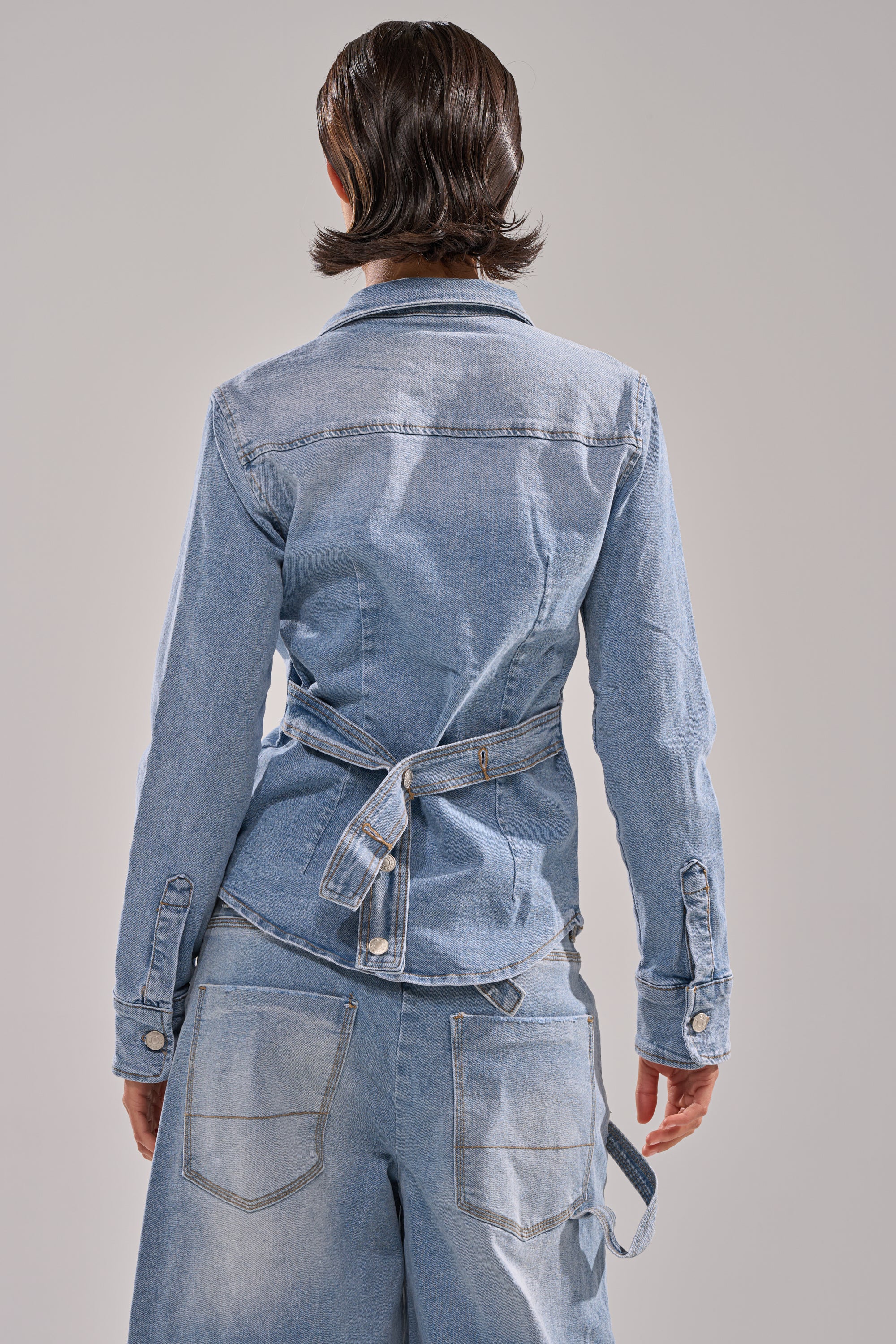 A person with short, dark hair stands with their back to the camera, wearing the ROCK MY WORLD DENIM BUTTON DOWN in light blue, belted at the back, paired with matching light blue jeans.