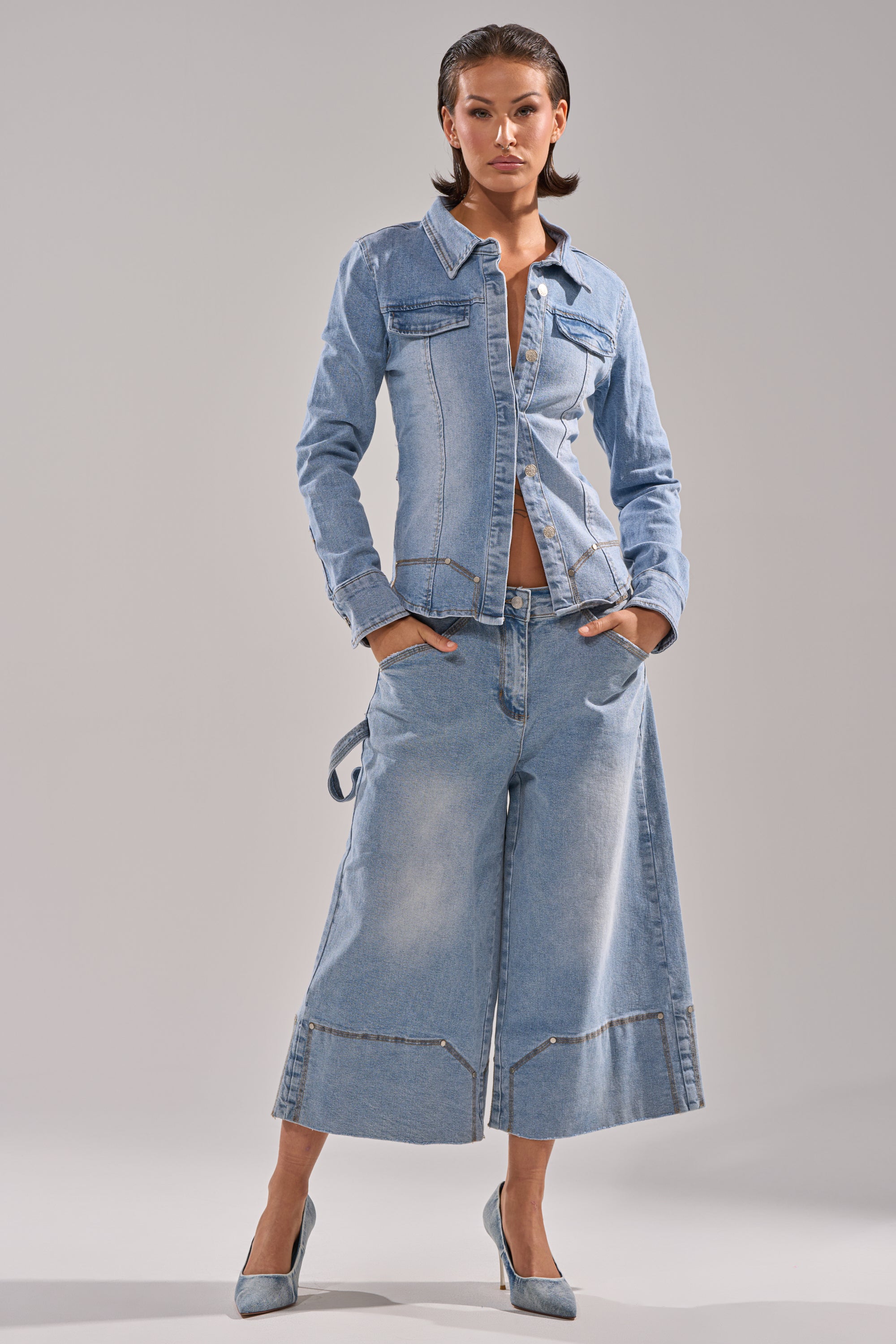 A woman stands confidently in the ROCK MY WORLD DENIM BUTTON DOWN with matching wide-leg cropped jeans and blue heels against a plain, light grey background.