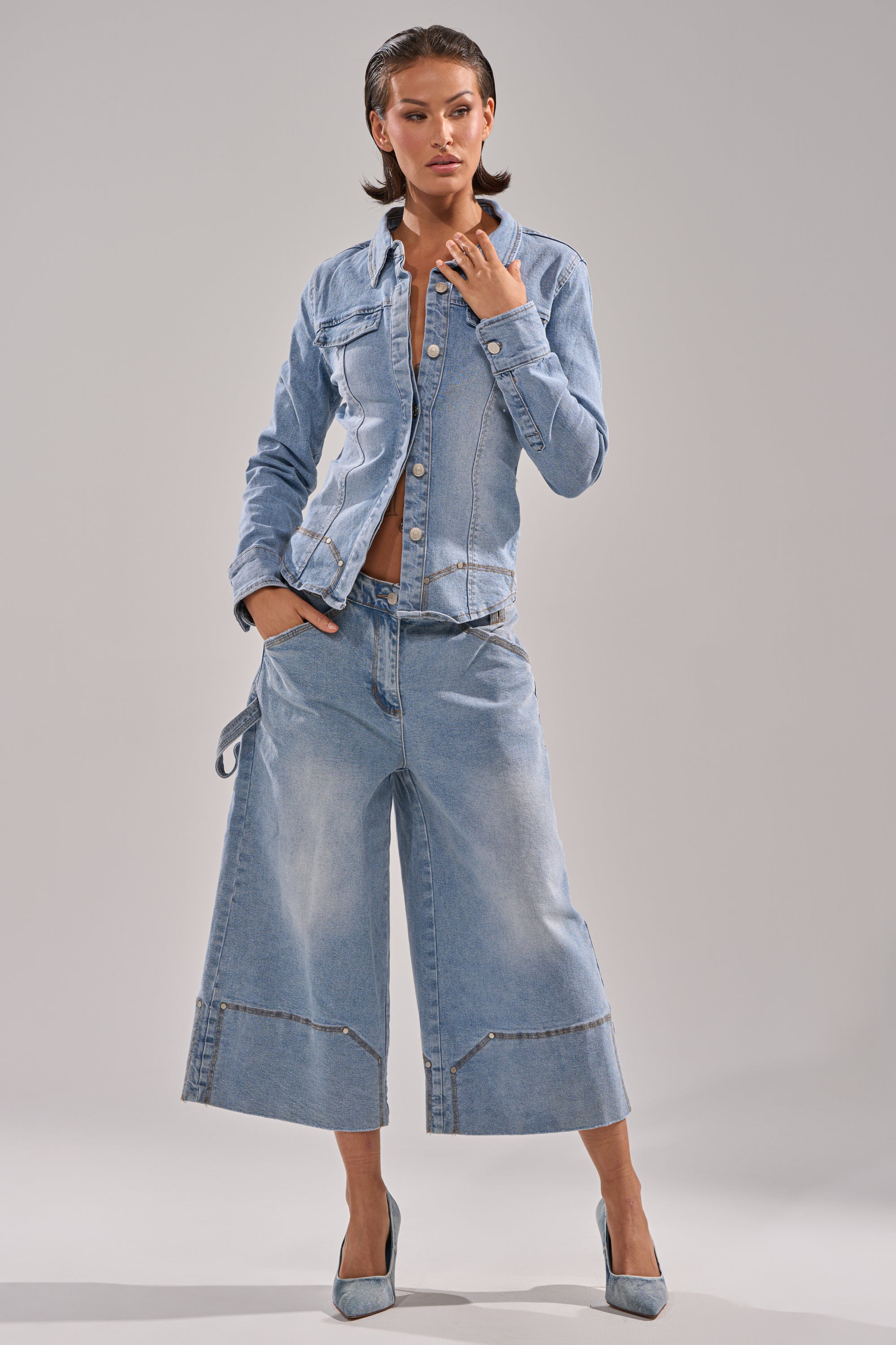 A woman poses in the light blue ROCK MY WORLD DENIM BUTTON DOWN and matching wide-leg culotte jeans with frayed hems, paired with denim high heels, against a plain gray background.