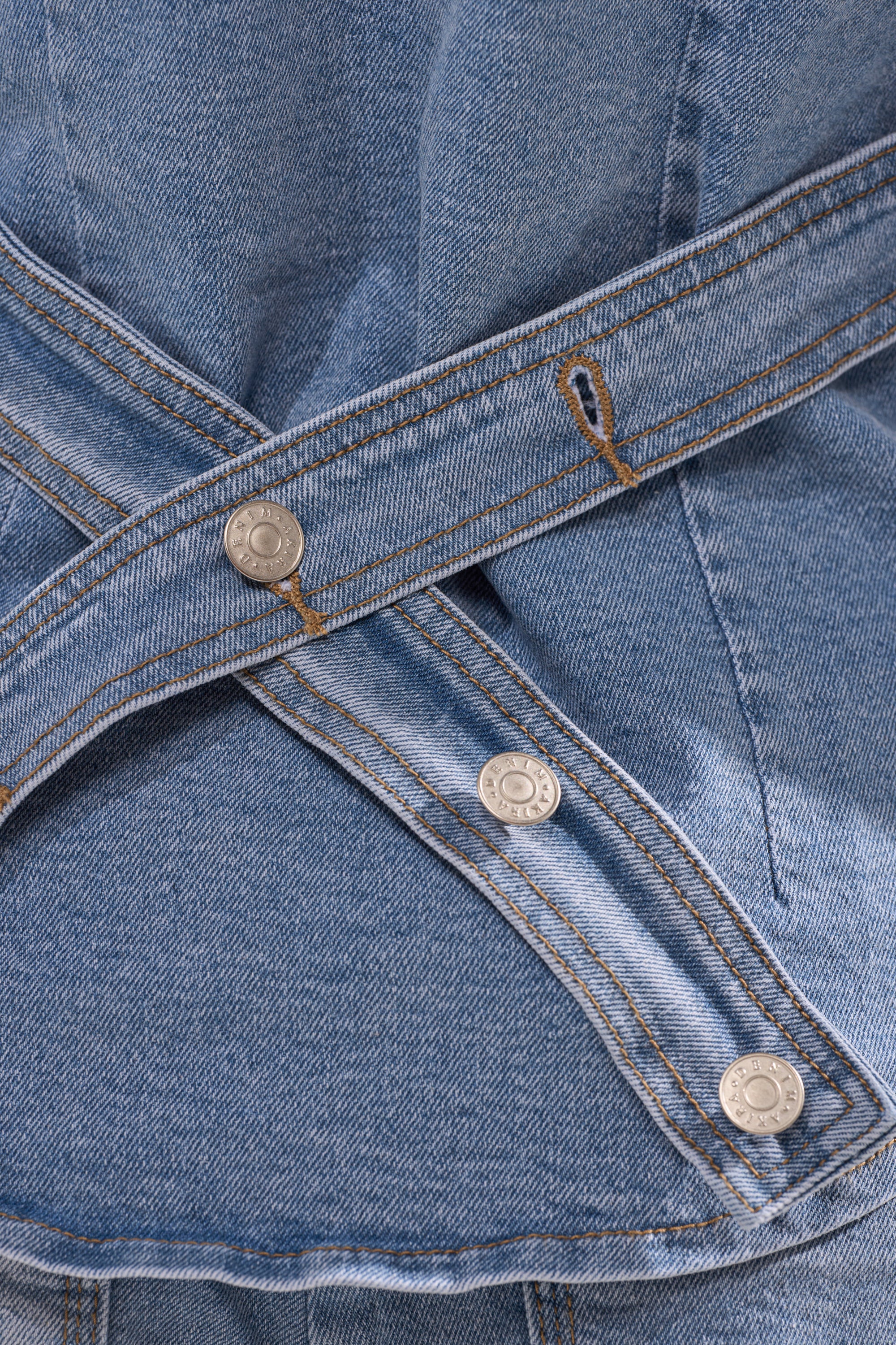 Close-up of blue denim fabric from the ROCK MY WORLD DENIM BUTTON DOWN, showcasing two overlapping straps with metallic snap buttons and visible brown stitching.