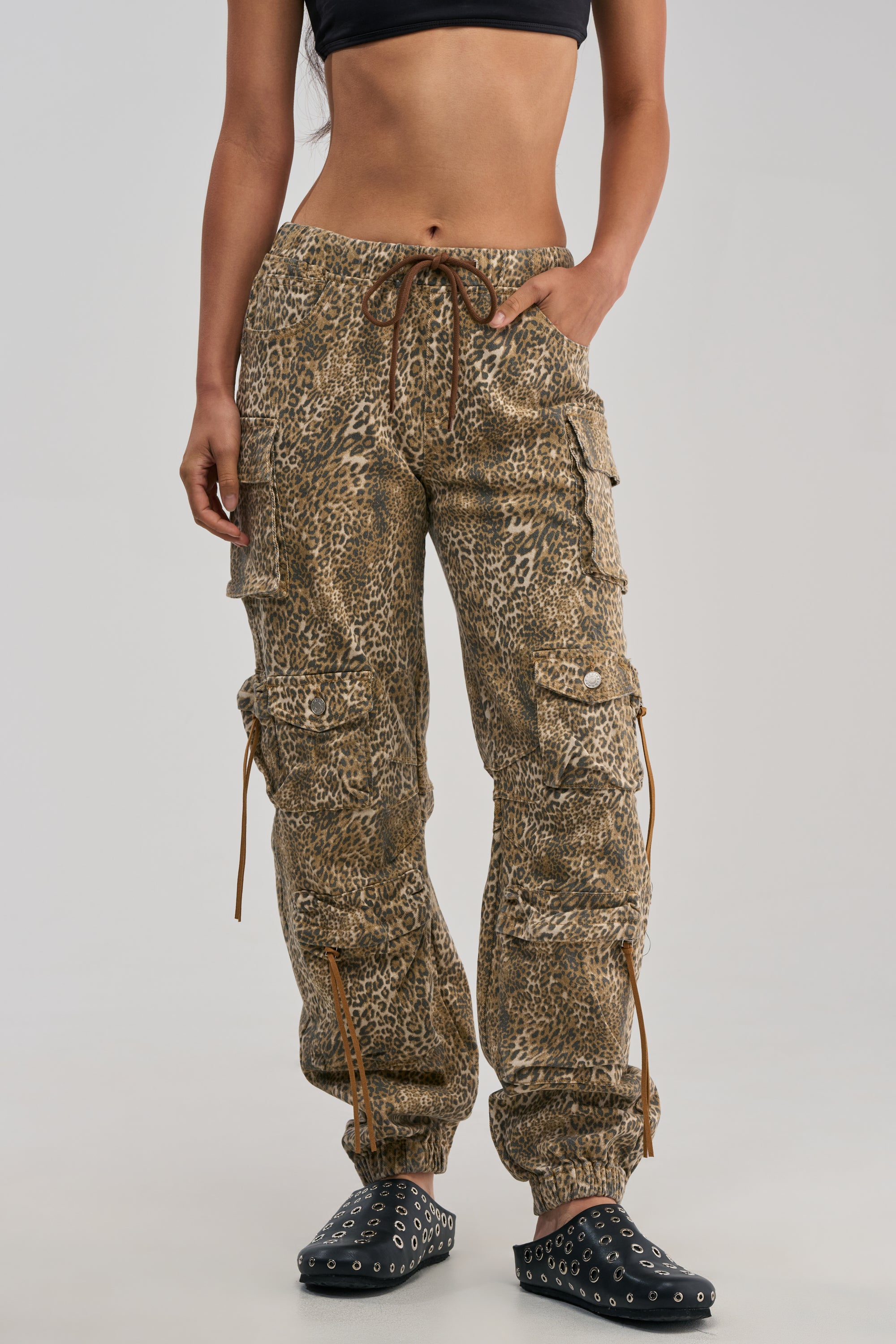 A person wearing a black crop top, the CHEETAH GIRL CHEETAH SISTA CARGO JOGGER pants with drawstrings in a brown cheetah print, and black studded shoes stands against a plain background.