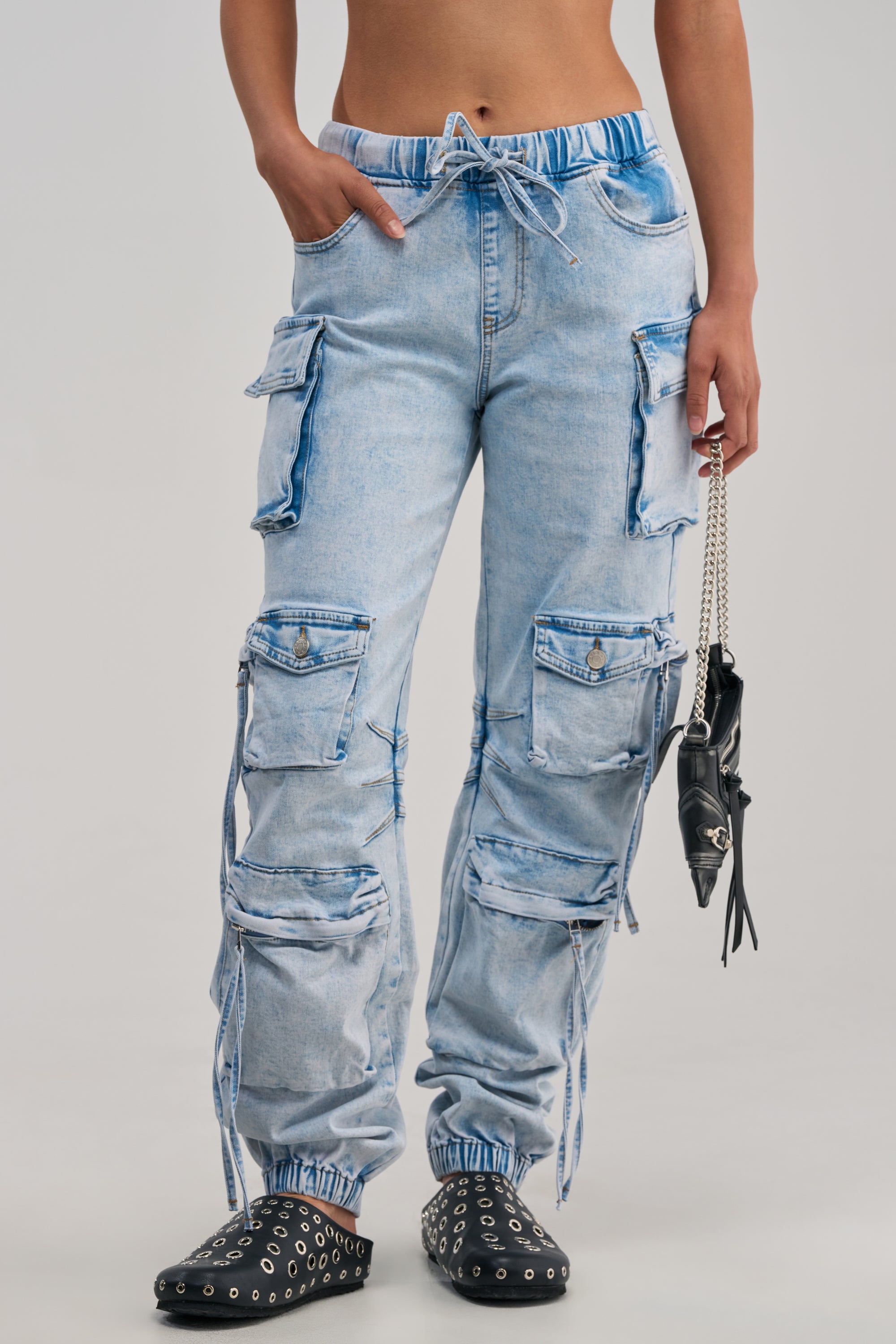 A person is wearing the CASUAL FOR DAYS DENIM JOGGER—light-wash streetwear pants featuring multiple large cargo pockets and drawstrings—paired with black studded shoes and holding a small black handbag. The background is plain and light gray.