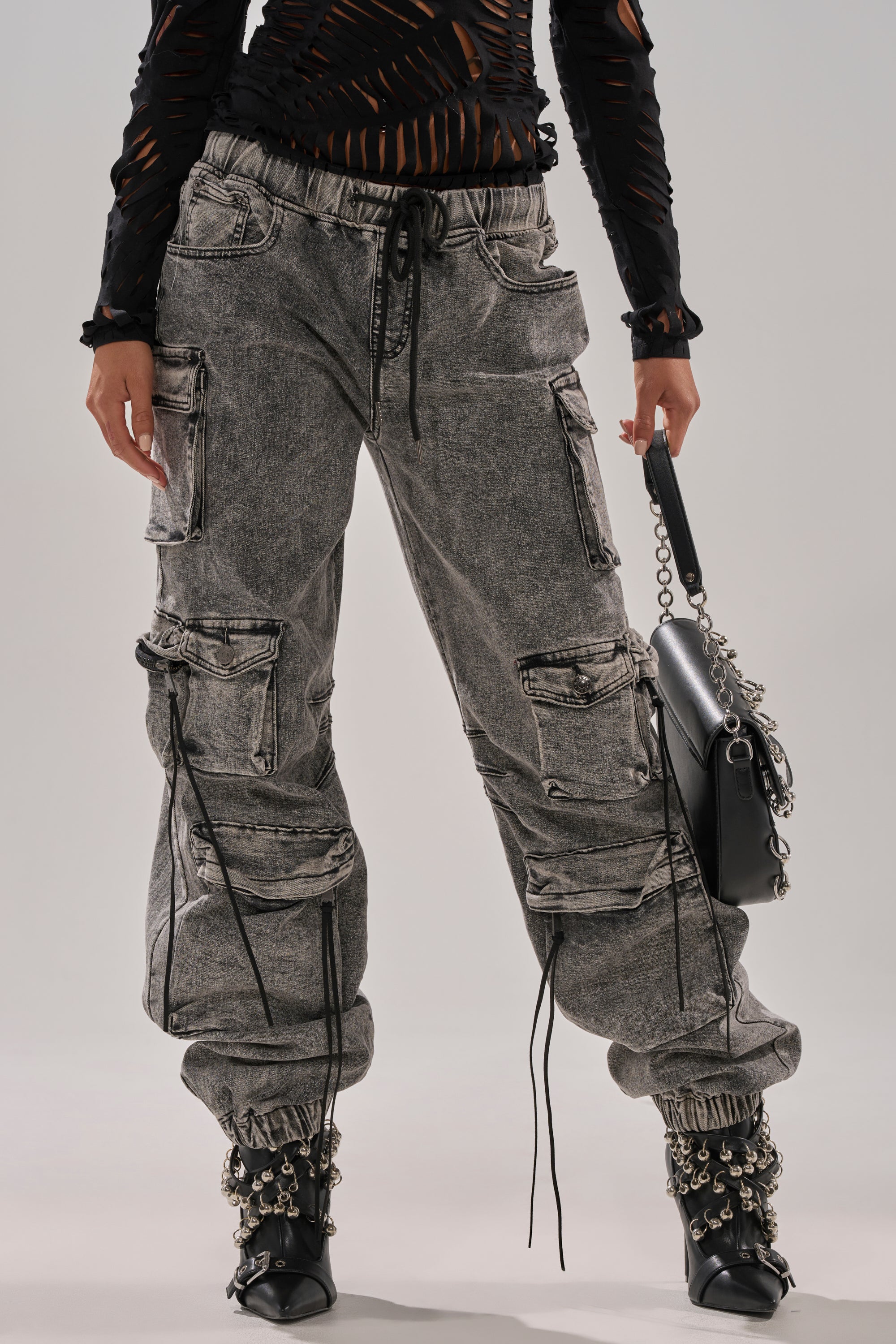 A person wears the CARGO FOR DAYS DENIM JOGGER—streetwear-inspired gray cargo pants with zippers and drawstrings—paired with a black cutout long-sleeve top, black chain-detailed boots, and a black handbag featuring a chain strap.