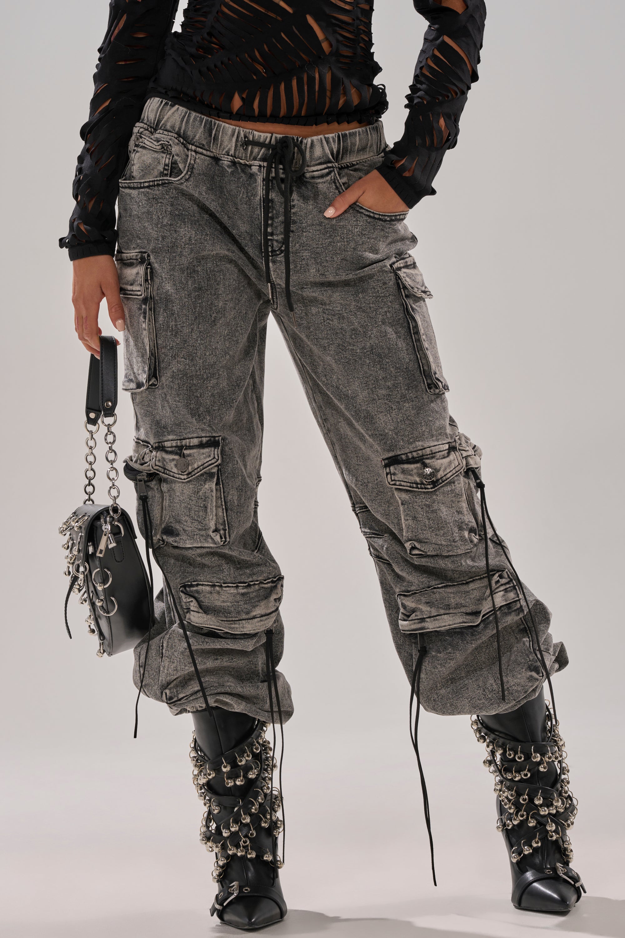 Someone wearing the CARGO FOR DAYS DENIM JOGGER—distressed black streetwear cargo pants with zippers and drawstrings—paired with a ripped black long-sleeve top, chain-detailed black boots, and a small black bag with metallic accents.