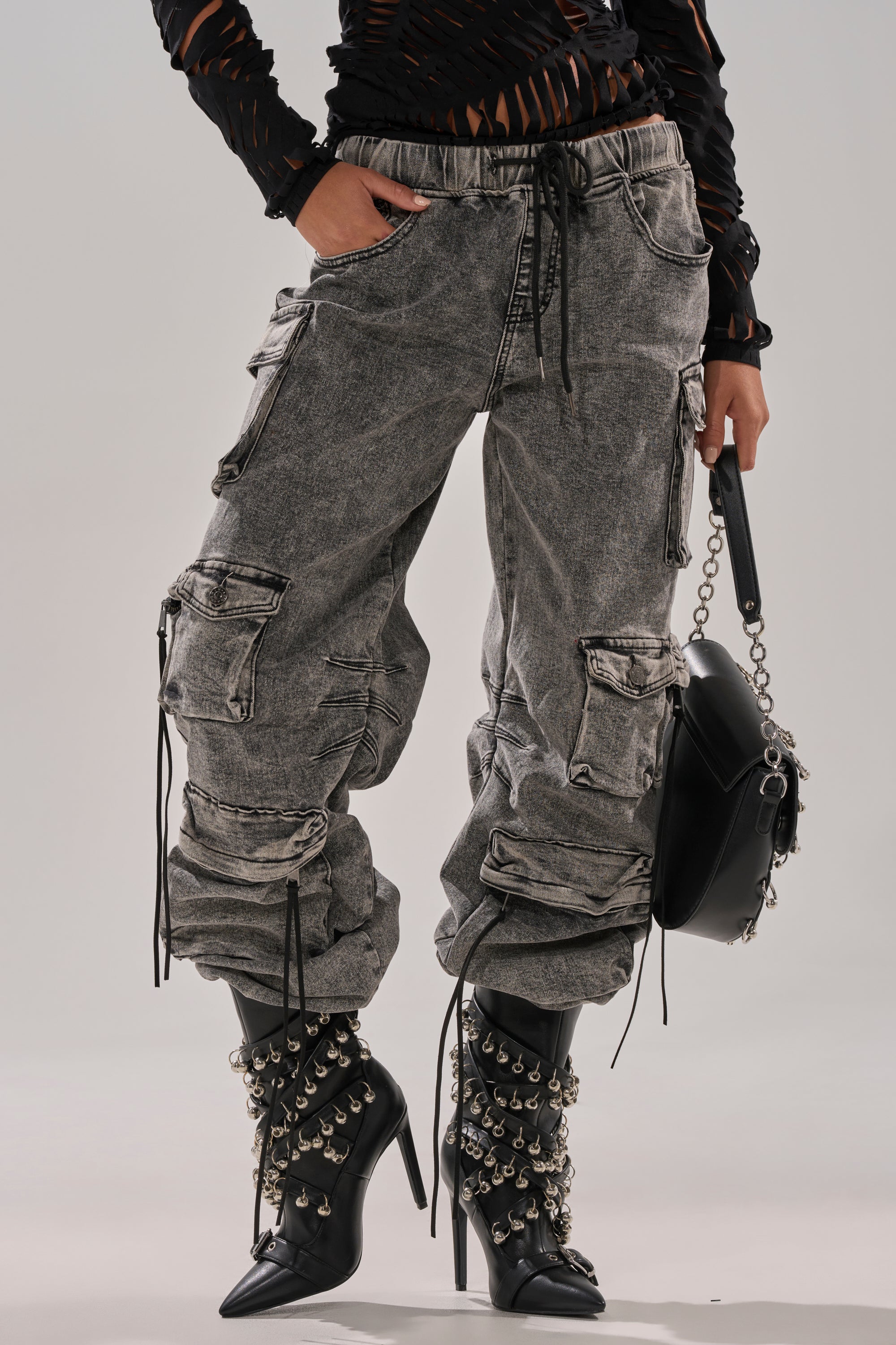 Wearing the CARGO FOR DAYS DENIM JOGGER in acid-wash gray with drawstrings, a ripped black top, studded black high-heeled boots, and holding a black chain-strap handbag.