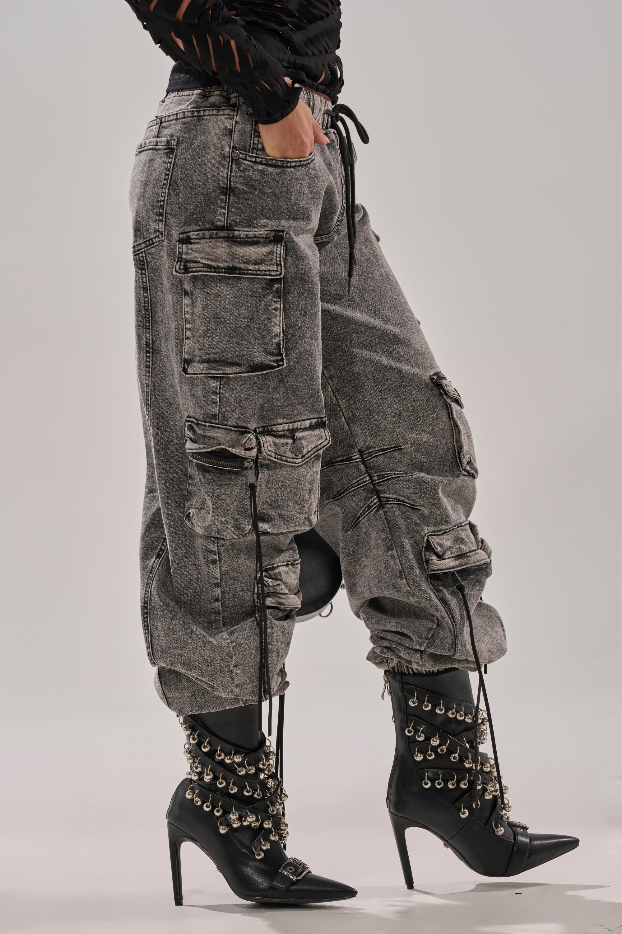 Wearing the CARGO FOR DAYS DENIM JOGGER—loose, faded gray cargo denim pants with multiple pockets—paired with black high-heeled boots with metal studs and chains, standing on a light gray background.
