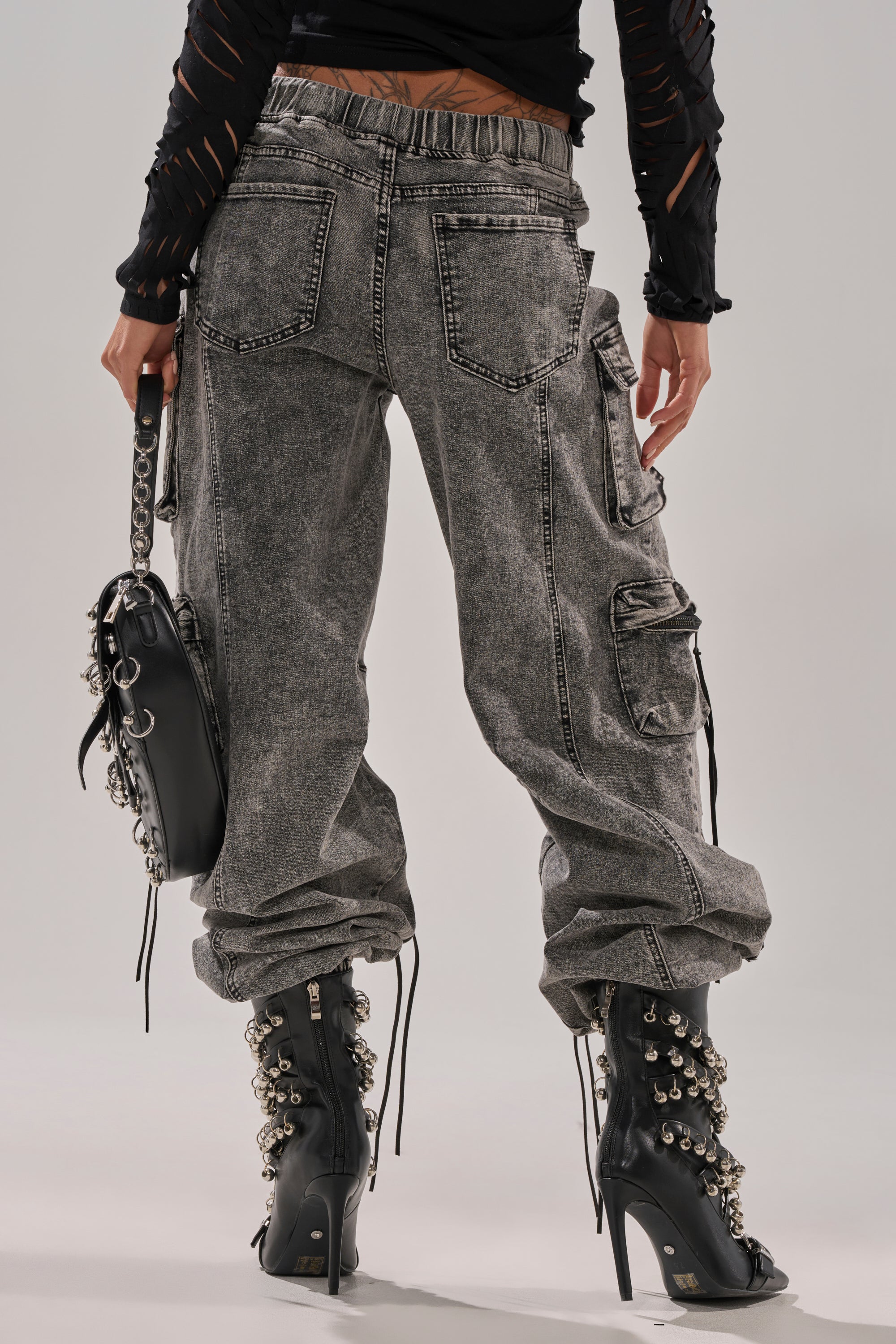 Wearing the CARGO FOR DAYS DENIM JOGGER, black lace-up high-heeled boots with metal rings, and holding a black chain-detail handbag, a person stands with their back to the camera.