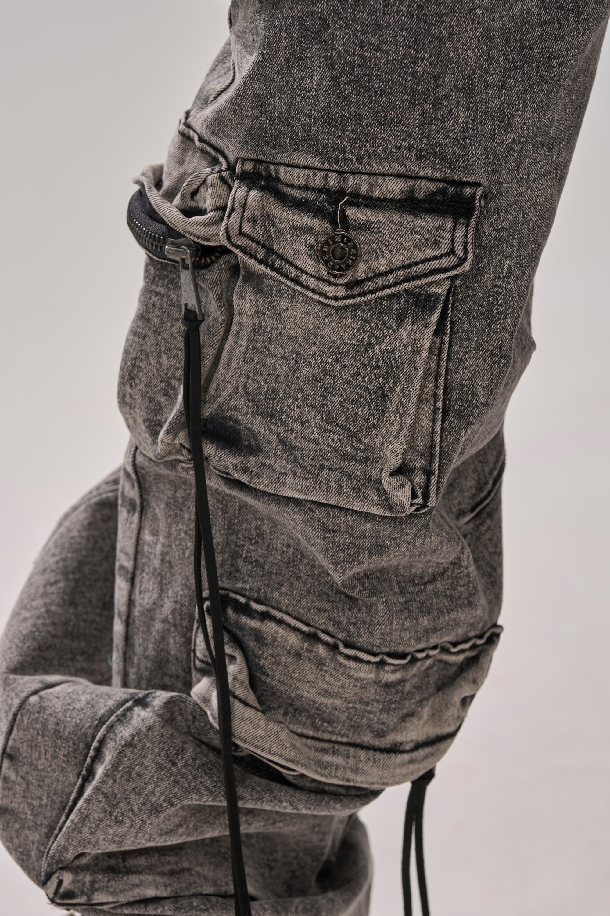 Close-up of the CARGO FOR DAYS DENIM JOGGER, gray acid-washed cargo pants with flap pockets, black drawstrings, detailed stitching, and textured fabric for a modern streetwear look.