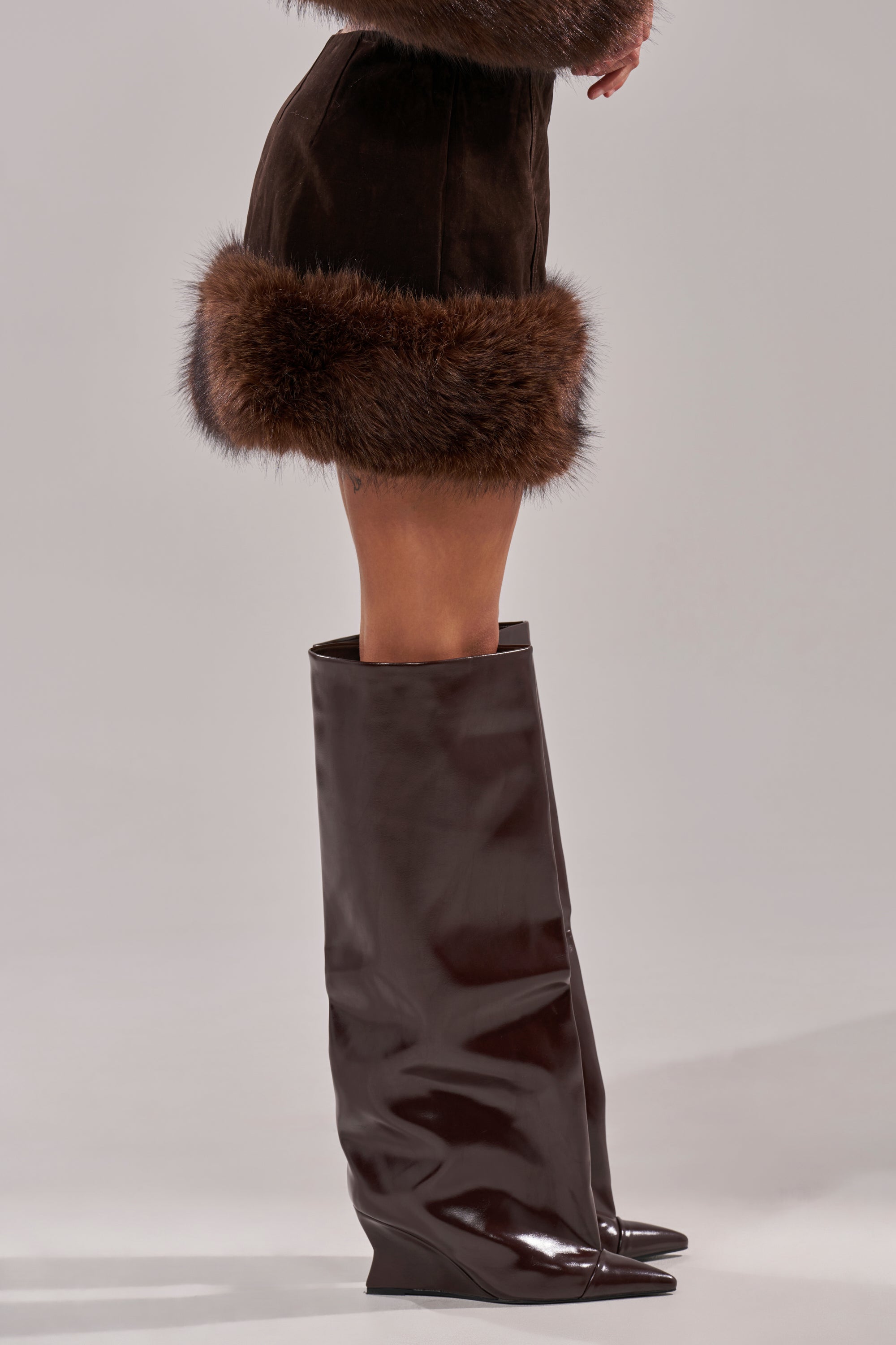 A person models the WILD SIDE FUR TRIM MINI SKIRT in dark brown with tall, glossy, over-the-knee dark brown boots, posed before a plain light gray background. 3