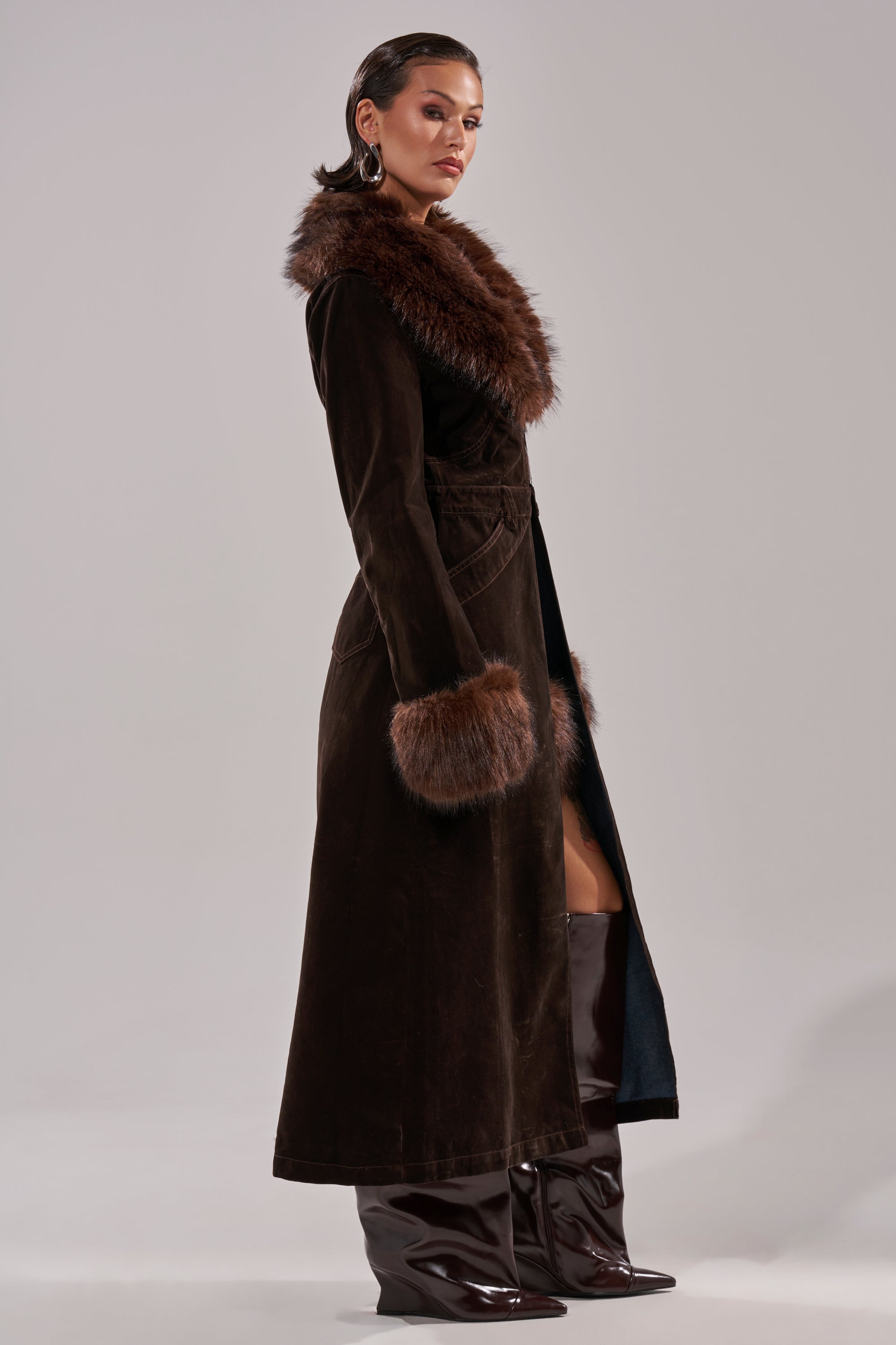 一位身穿 WILD SIDE 天鹅绒牛仔风衣的女士，风衣饰有大胆的人造毛皮领口和袖口，搭配闪亮的过膝长靴，站在纯色浅色背景前，自信地侧目而视。