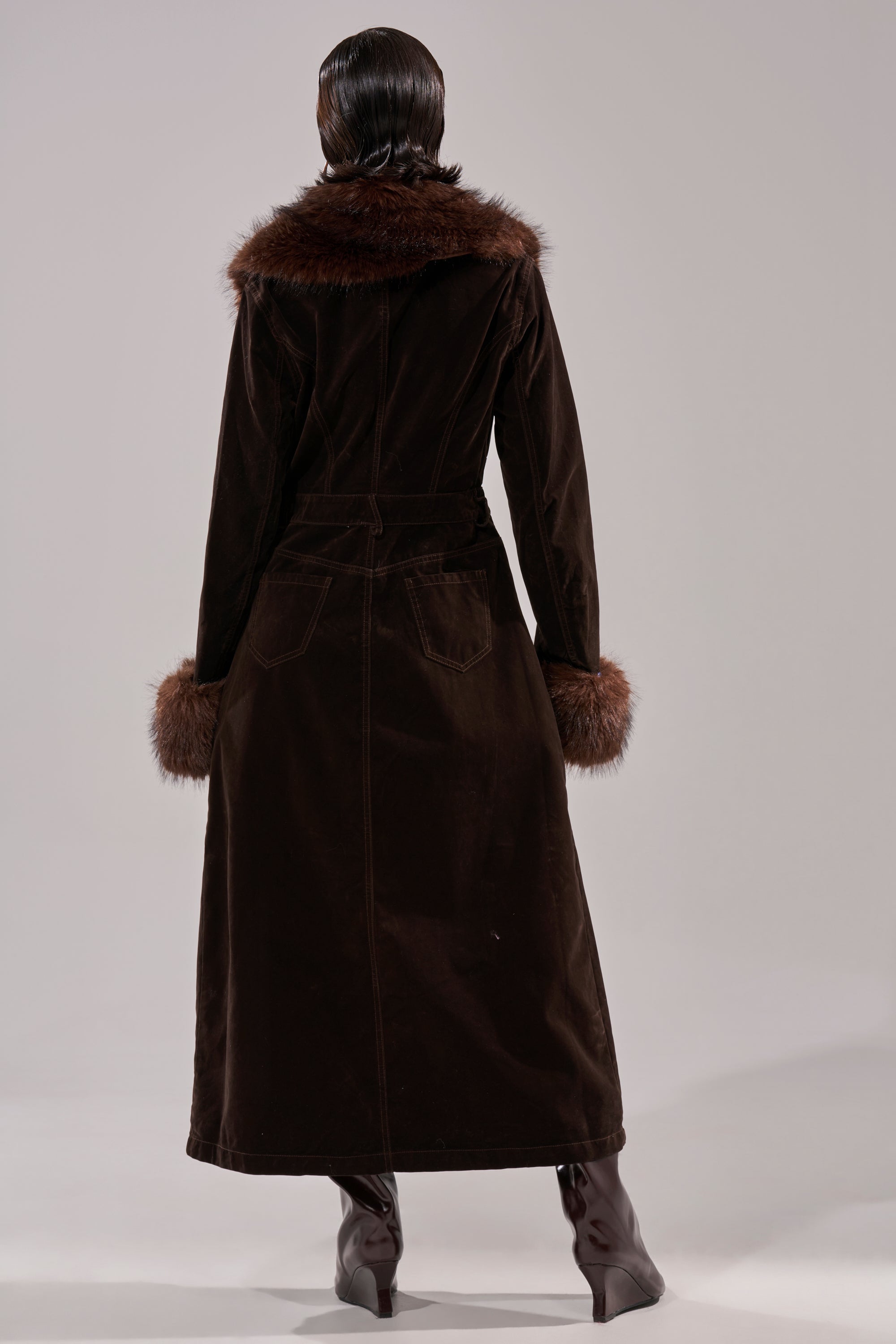 一个人背对着镜头，身穿WILD SIDE天鹅绒牛仔风衣，领口和袖口饰有人造毛皮，脚蹬棕色高跟靴，深色头发梳得一丝不苟。背景简洁浅淡。