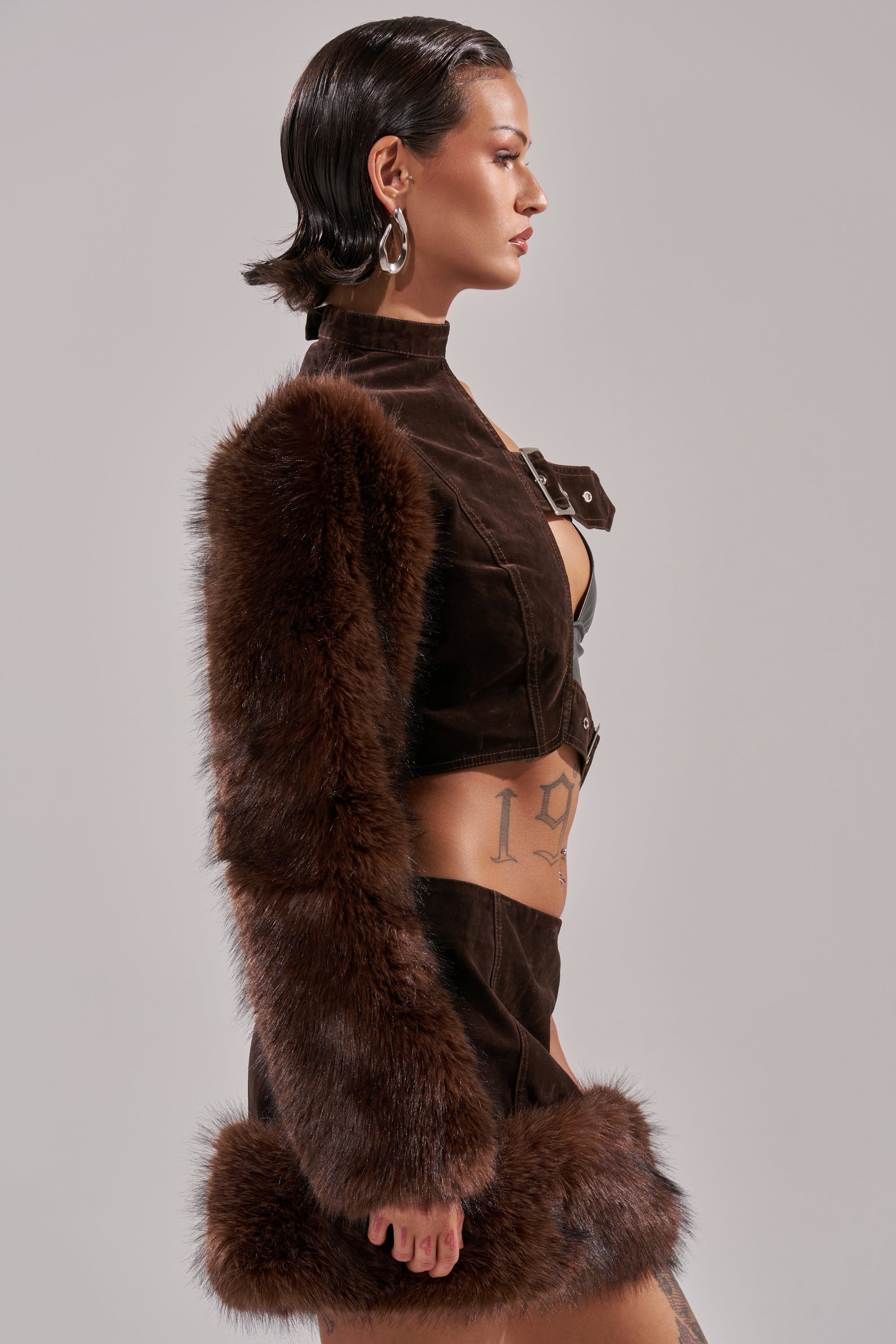 A woman with slicked-back dark hair wears the WILD SIDE SINGLE SLEEVE FUR TOP and matching skirt, styled with hoop earrings and visible side tattoos, standing in profile against a neutral background. 1