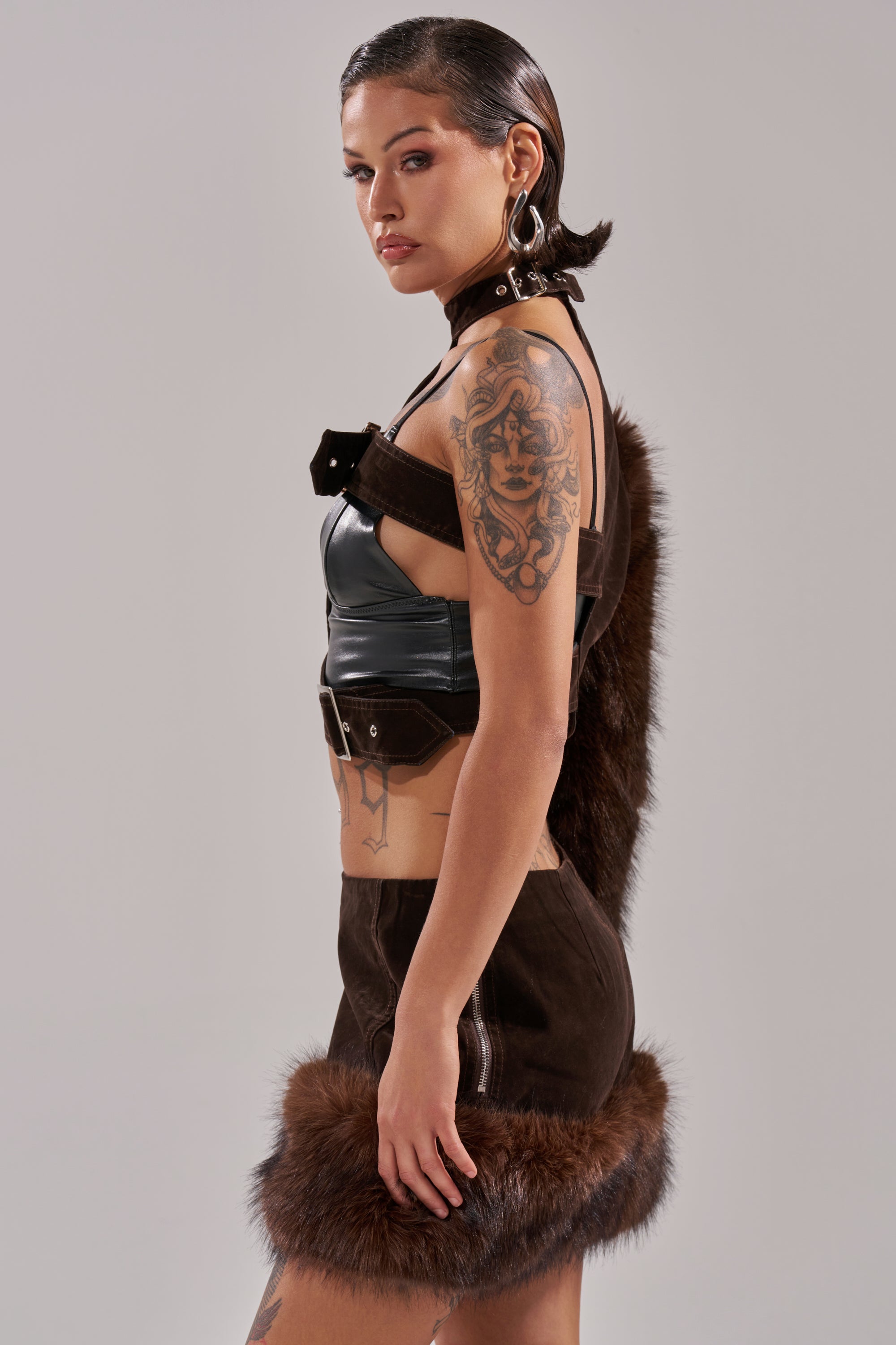 A tattooed woman poses in profile against a plain light gray background, wearing the WILD SIDE SINGLE SLEEVE FUR TOP—black leather with brown fur details—a matching brown skirt with fur trim, and large silver hoop earrings. 2