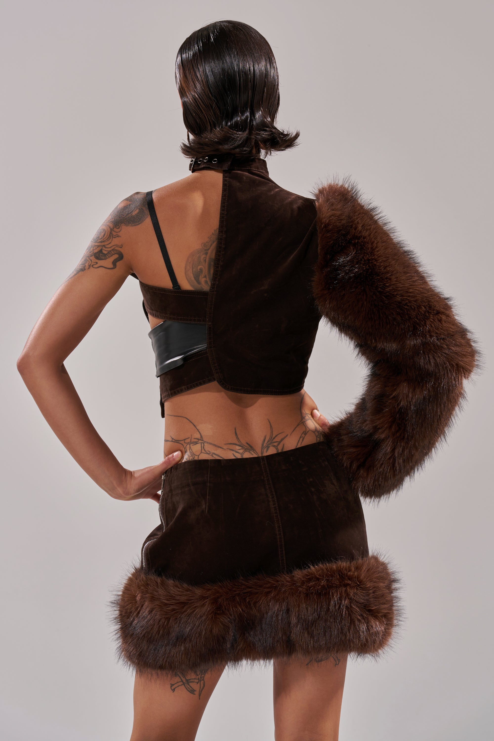 Viewed from behind against a plain light grey background, a person with short, slicked-back hair showcases tattoos while wearing the WILD SIDE SINGLE SLEEVE FUR TOP in dark brown with faux fur sleeve and asymmetric design. 3
