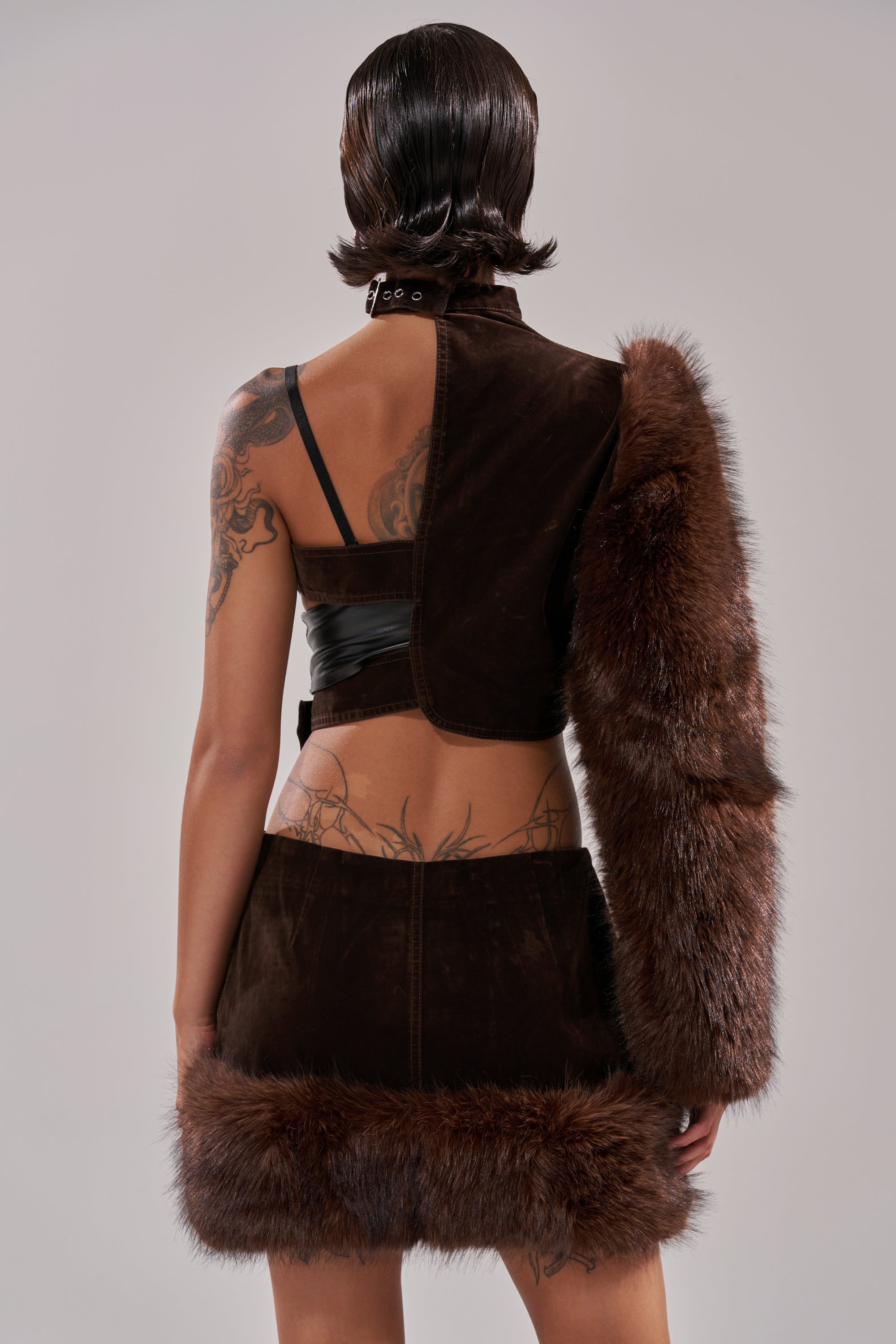 A person with short, slicked-back hair is seen from behind, wearing the WILD SIDE SINGLE SLEEVE FUR TOP with faux fur and an asymmetrical design that reveals back tattoos and a black strap. The background is a neutral gray. 4