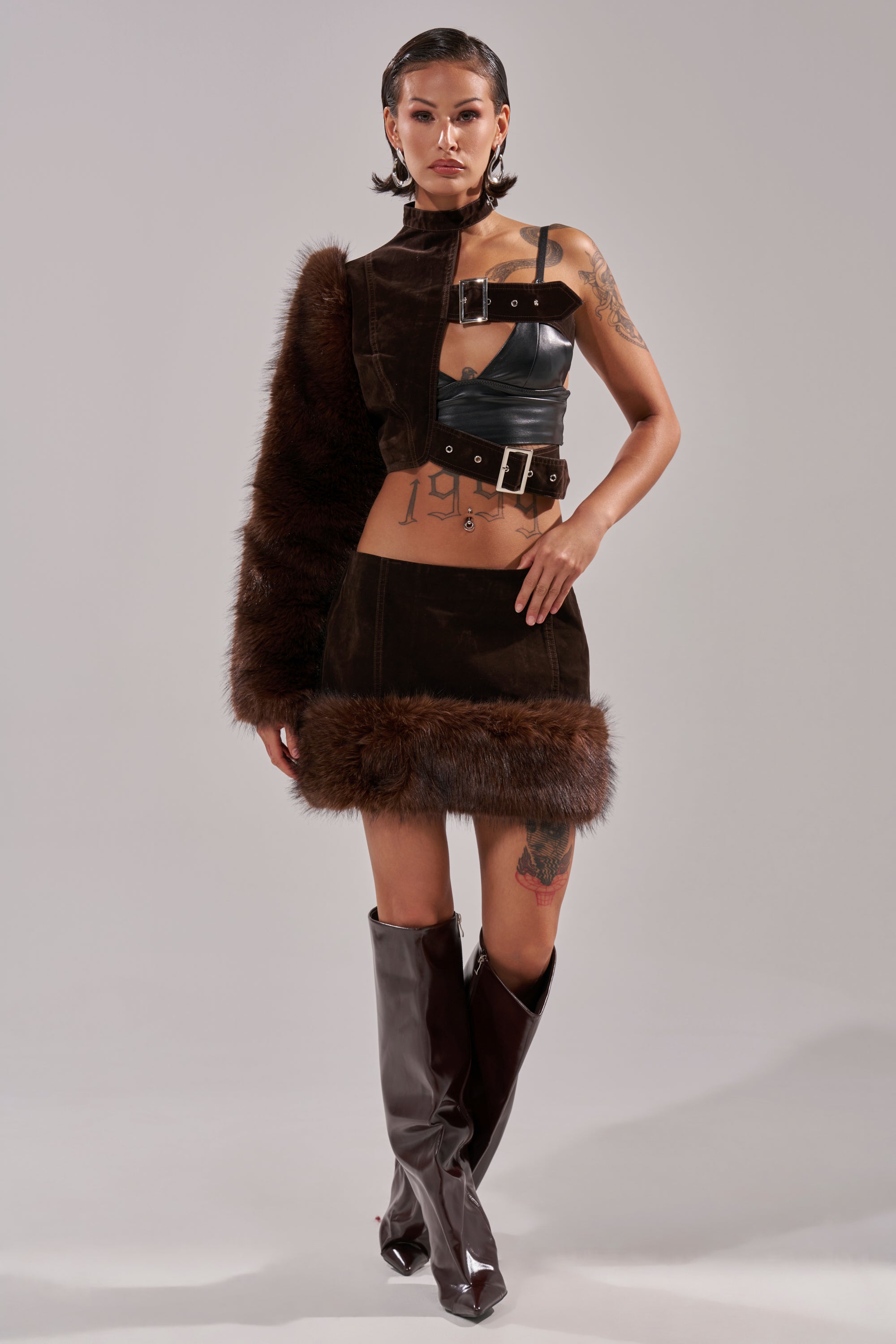 A model with tattoos and wet-look hair poses against a plain background wearing the WILD SIDE SINGLE SLEEVE FUR TOP, styled with a leather strap mini skirt and tall brown boots. 8