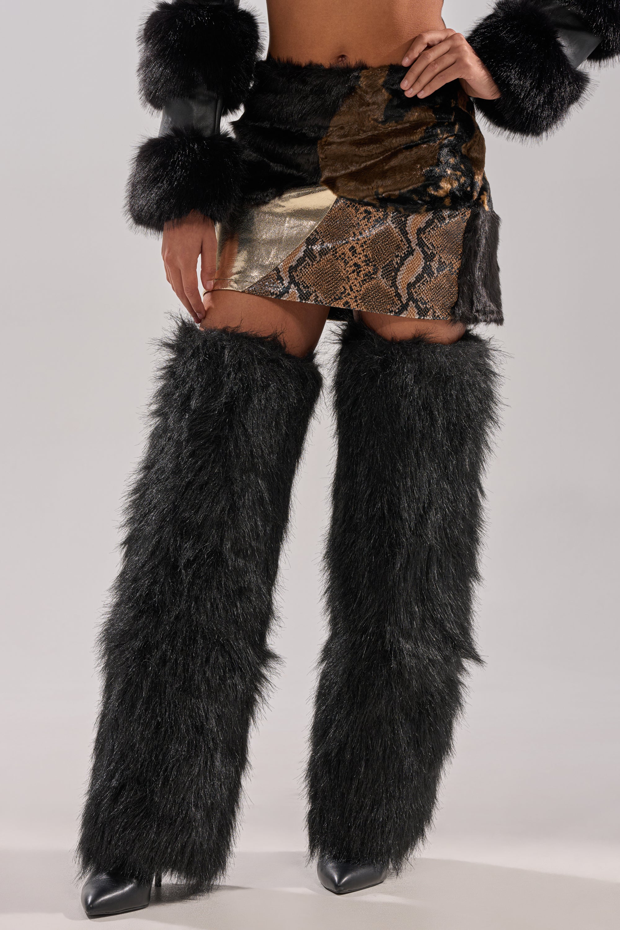 A person wears black faux fur leg warmers over pointed-toe heels, the ALWAYS WANT YOU BY MY SIDE ABSTRACT FUR MINI SKIRT with mixed textures, and a jacket with black faux fur cuffs. The upper body is cropped out of the image.