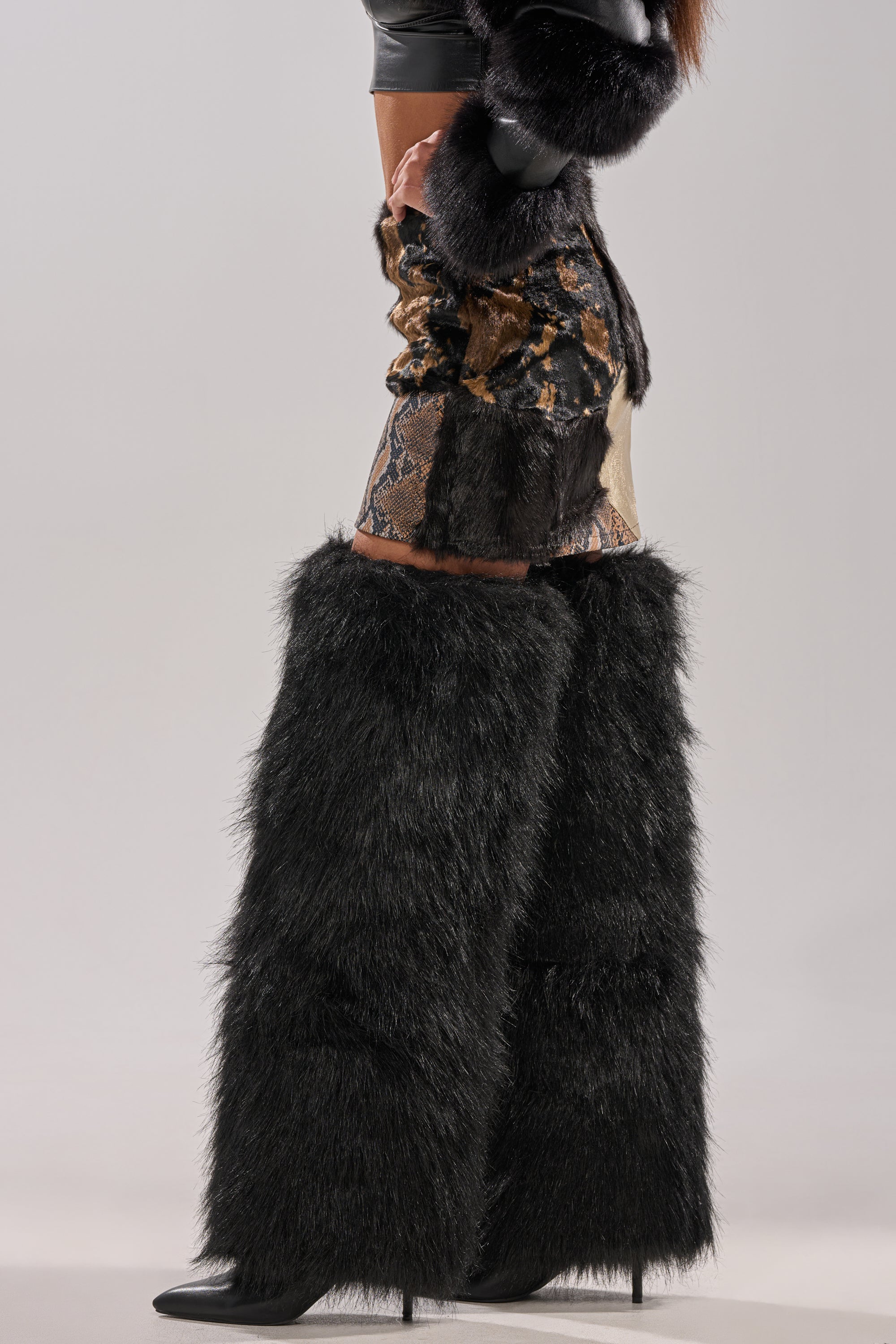 A person wearing the ALWAYS WANT YOU BY MY SIDE ABSTRACT FUR MINI SKIRT, large black furry pants over high heels, black gloves, and a black fur stole stands against a plain background.