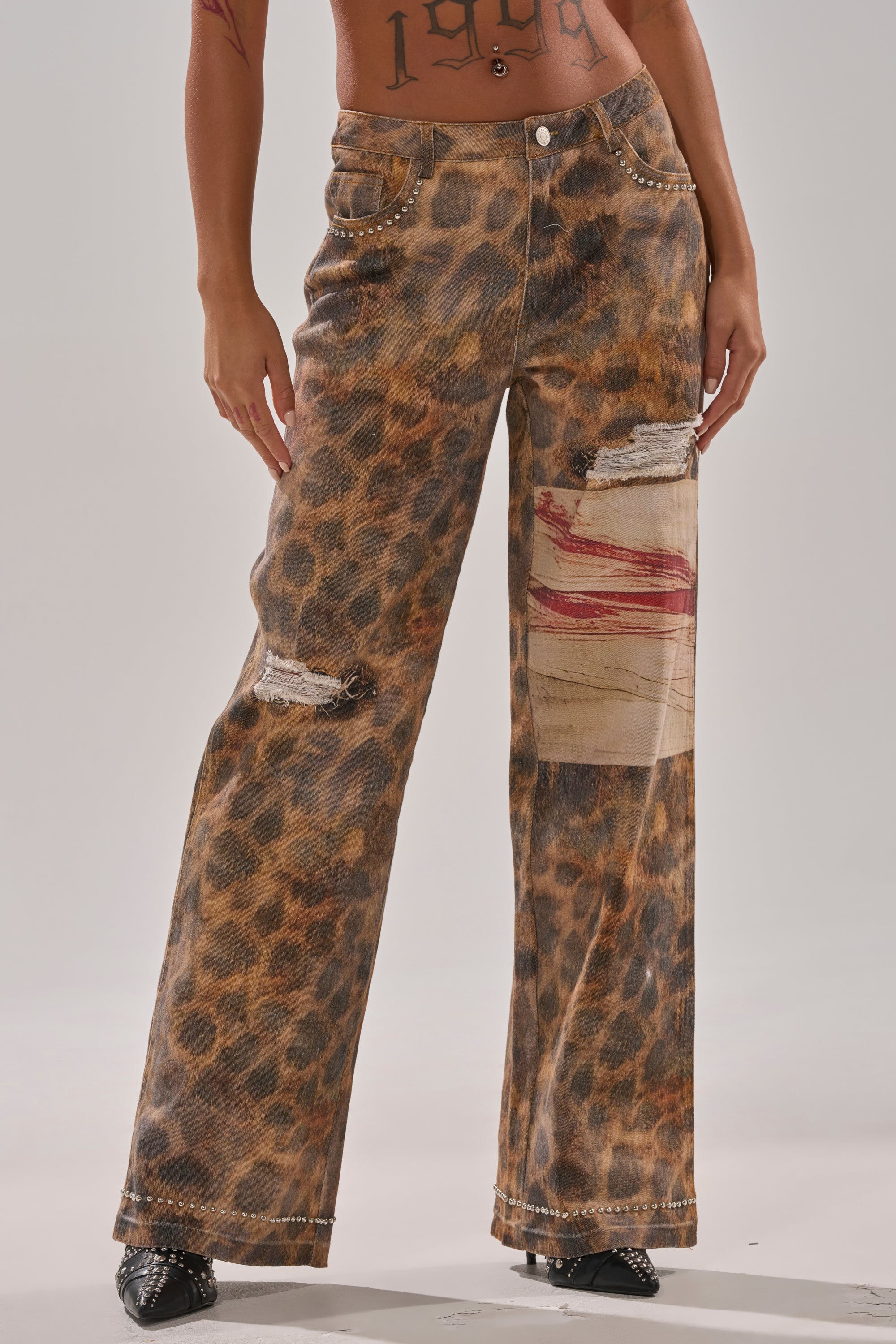 Person wearing BACK OF MY MIND WIDE LEG DENIM—high-rise leopard print jeans with a wide-leg cut, distressed details, patched rip featuring a red paint streak, and silver stud accents—standing against a plain background.