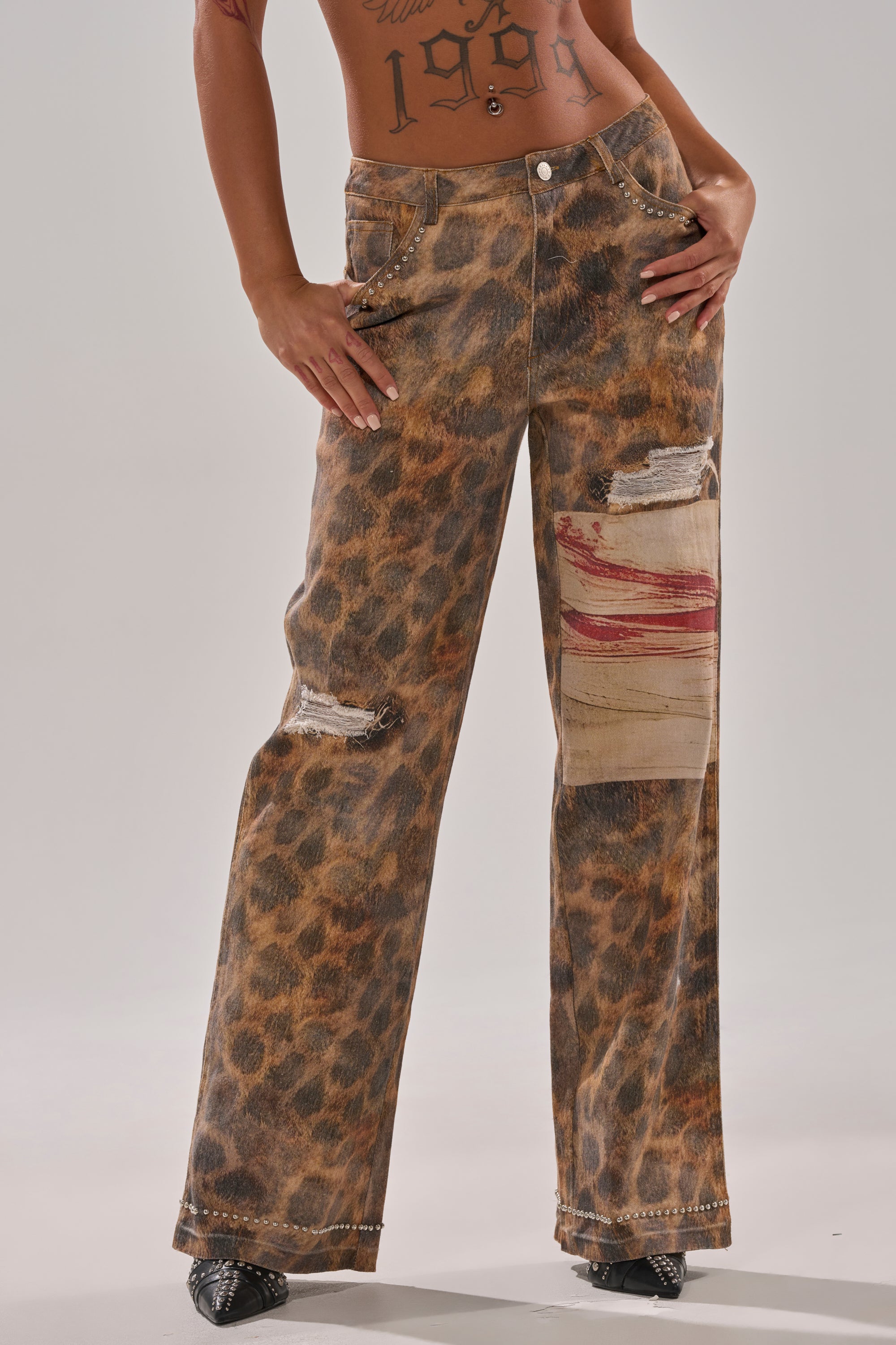 Someone wears BACK OF MY MIND WIDE LEG DENIM—high-rise, wide-leg jeans in a leopard print with ripped details and a beige patch with red streaks—standing against a plain background, tattoos visible on their torso.