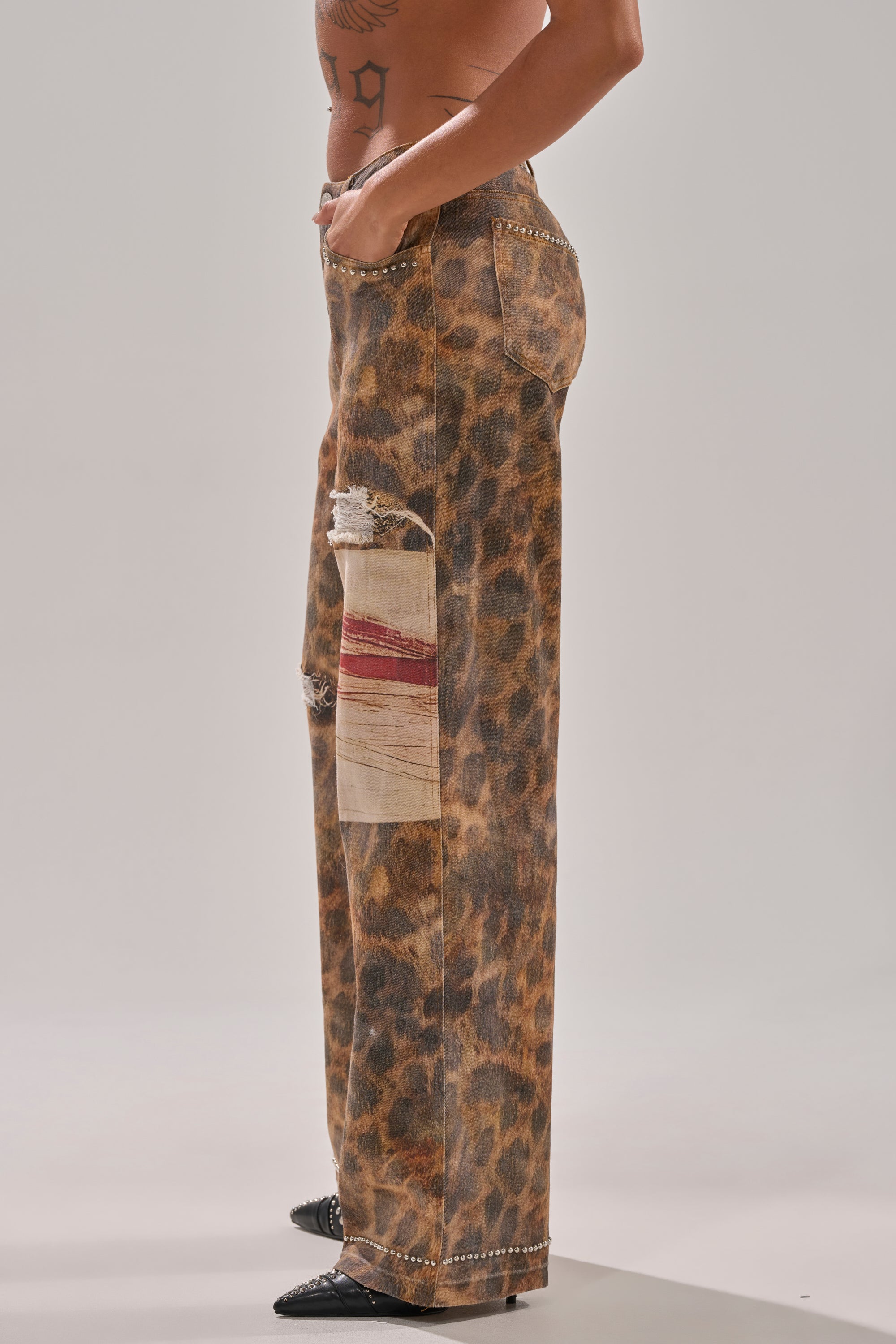 A person in BACK OF MY MIND WIDE LEG DENIM, featuring high-rise wide legs, leopard print, a large patch, and distressed thigh details, stands in profile against a plain background.