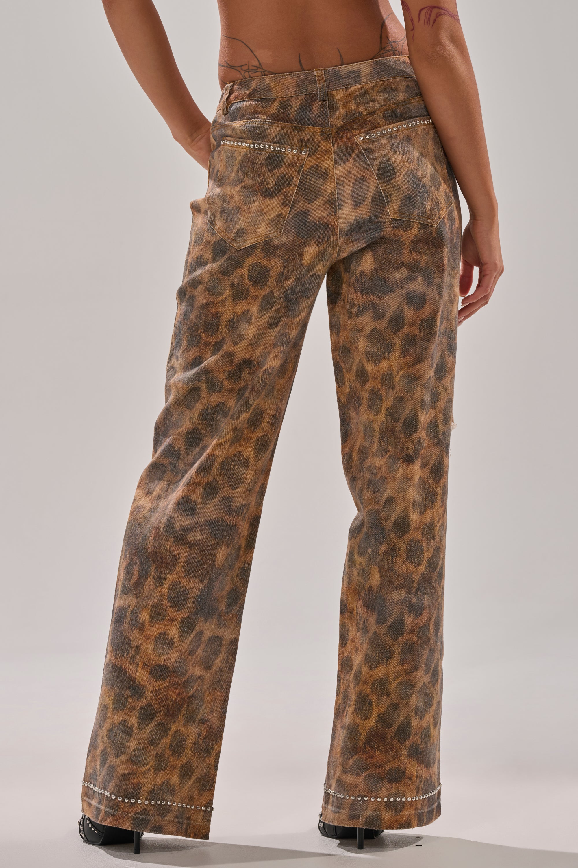 A person stands with their back to the camera, wearing BACK OF MY MIND WIDE LEG DENIM—high-rise, leopard-print wide-leg jeans with decorative stitching and metal studs on the pockets—against a plain, light background.