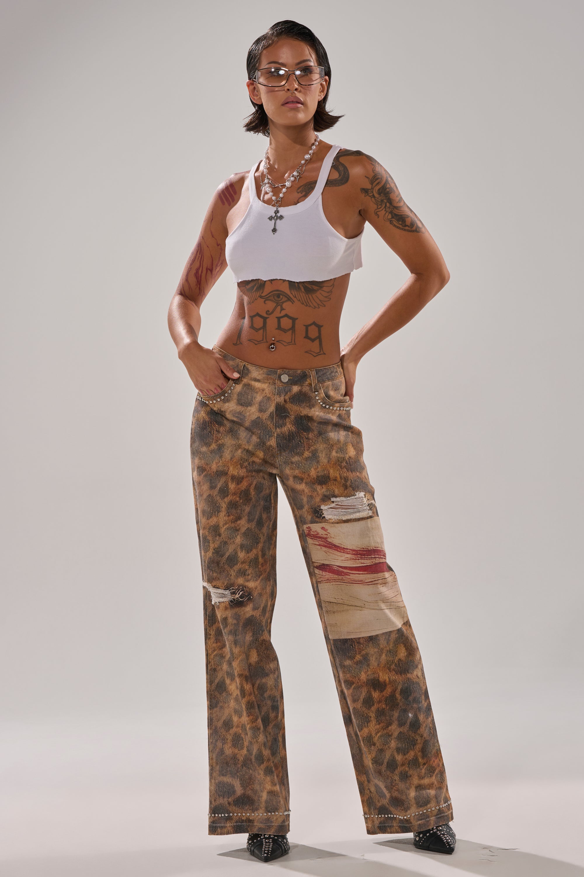 A person with short dark hair, glasses, and tattoos wears a white tank top and the BACK OF MY MIND WIDE LEG DENIM—high-rise leopard print jeans with patches and rips—standing confidently against a plain light background.
