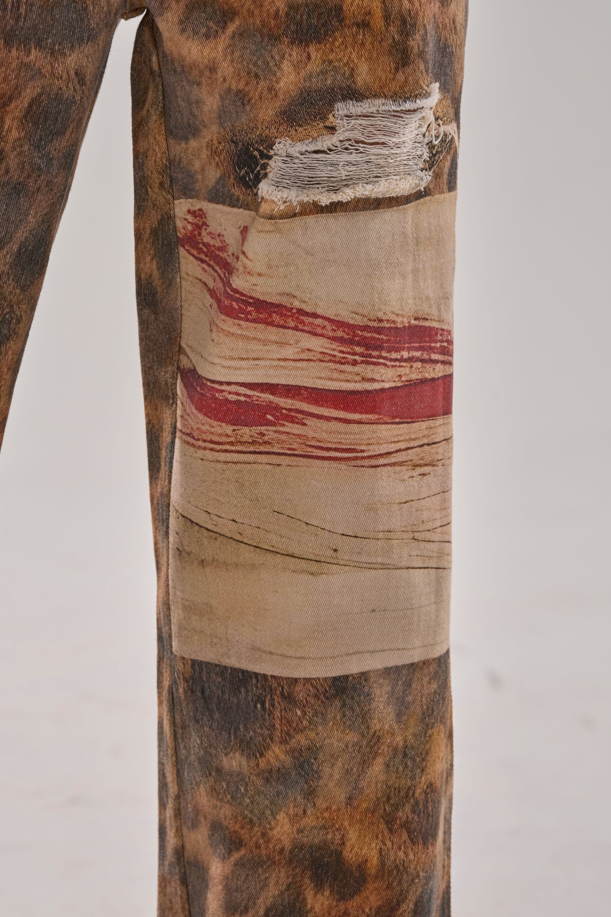 A close-up of BACK OF MY MIND WIDE LEG DENIM shows a pant leg with a bold leopard print, frayed patch, and a rectangular beige patch featuring red paint-like streaks.