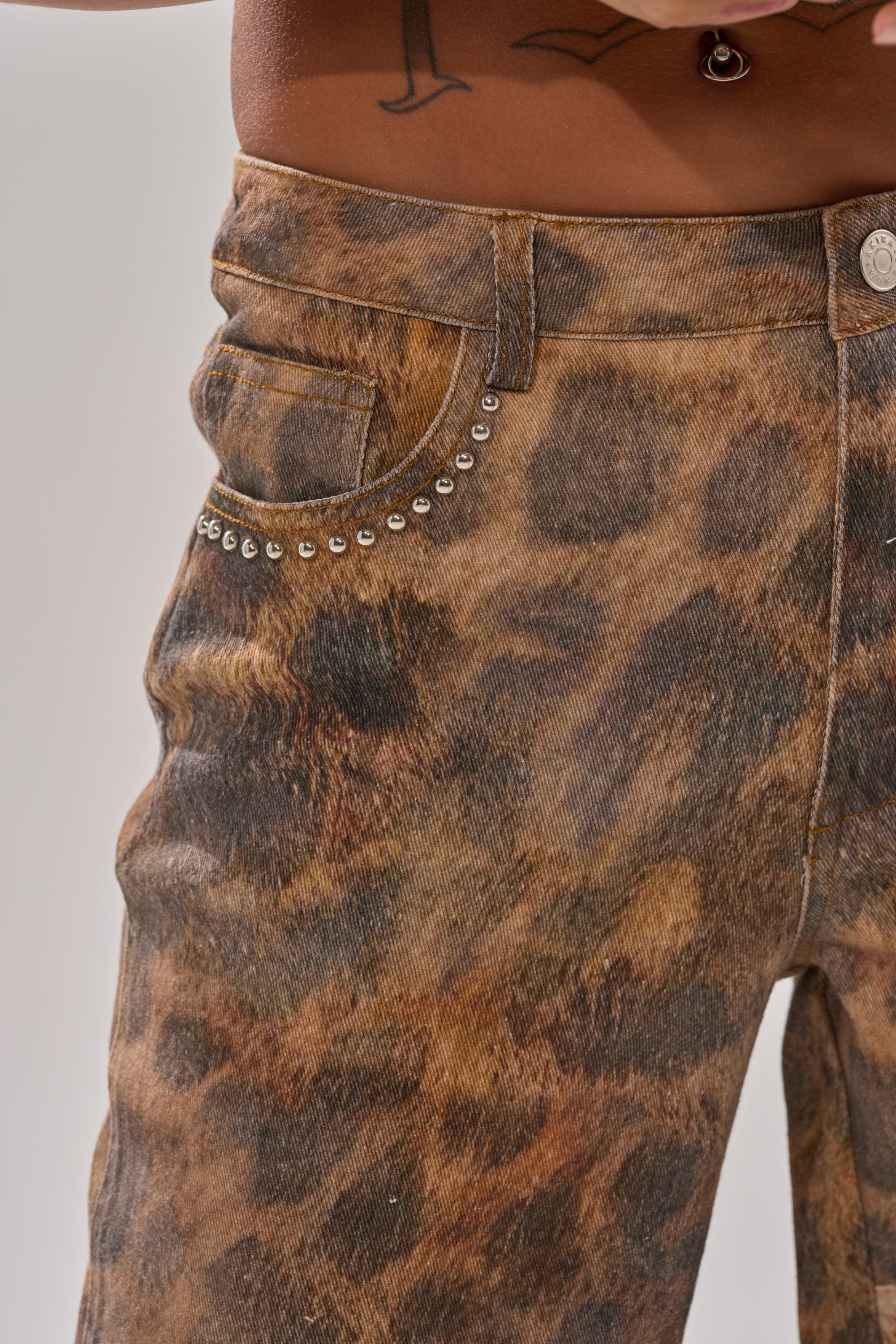 Close-up of a person wearing BACK OF MY MIND WIDE LEG DENIM—high-rise, brown leopard print jeans with silver studs on the front pocket. A tattoo is visible on their lower torso, and no shirt is shown.