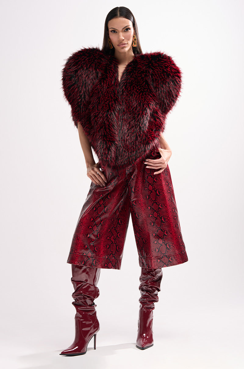A woman poses confidently in the EXTRA IS MY MIDDLE NAME SNAKE BERMUDA SHORT, paired with a red and black faux fur jacket and shiny red knee-high boots, against a plain white background.