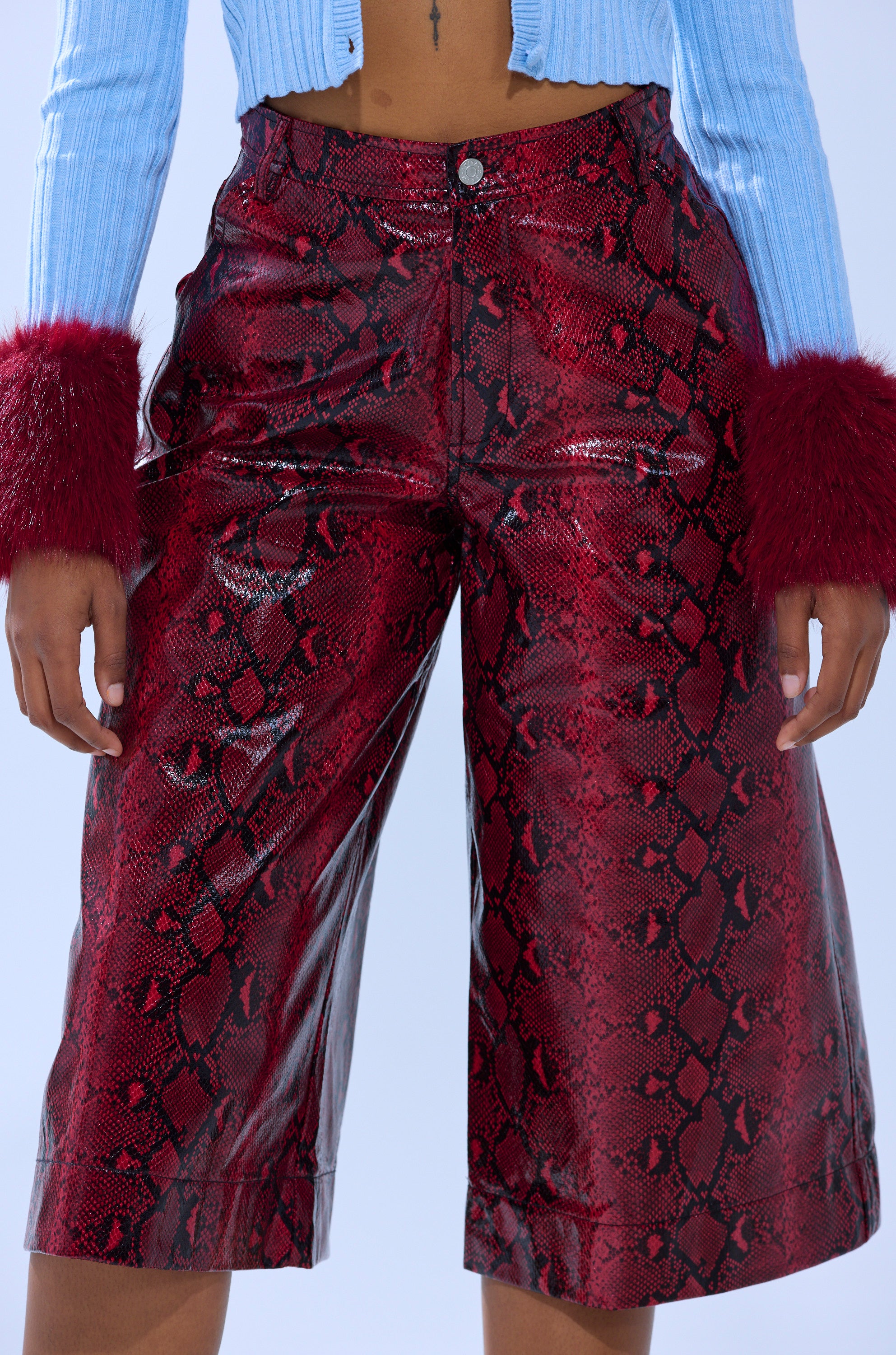 A person wears the EXTRA IS MY MIDDLE NAME SNAKE BERMUDA SHORT in shiny red embossed snakeskin print, paired with a light blue ribbed top and maroon faux fur sleeve cuffs. The photo is cropped from shoulders to knees.